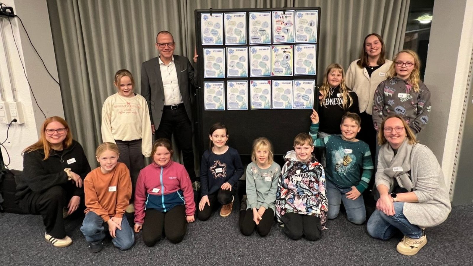 Gruppe von Kindern und Erwachsenen, die vor einem großen Plakat mit Zeichnungen und Texten stehen.