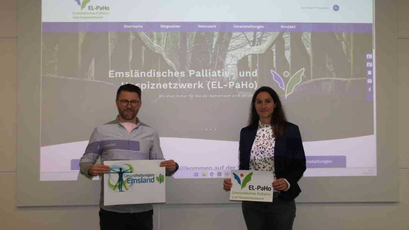 Zwei Personen halten Schilder mit den Logos von EL-PaHo und Gesundheitsregion Emsland vor einem Bildschirm mit einer Website.