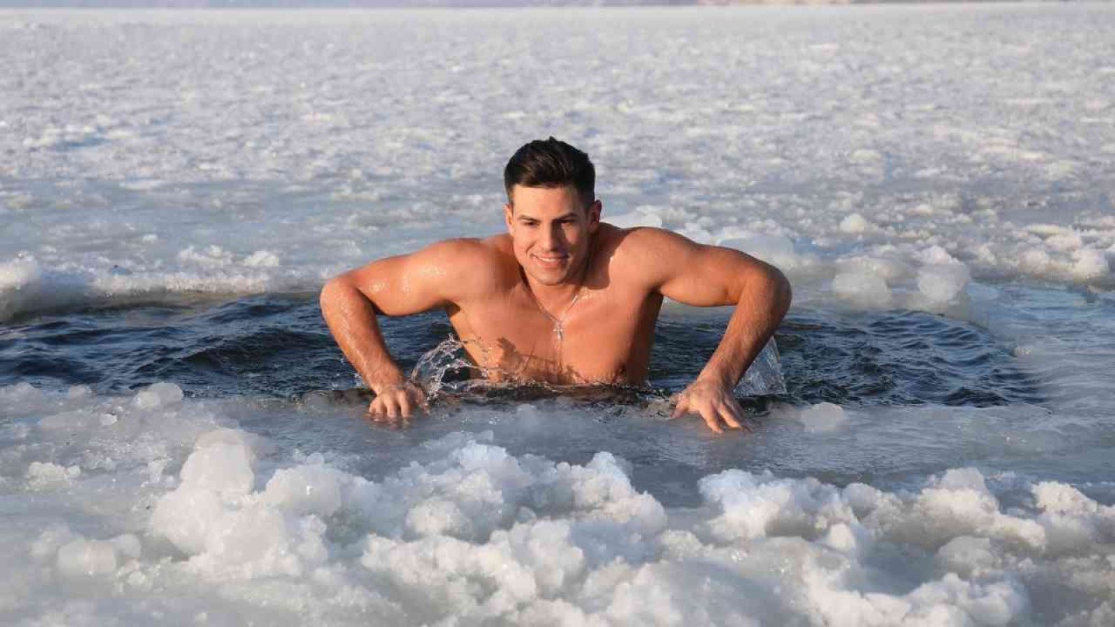 Mann, der aus einem Loch im Eiswasser auftaucht, mit muskulösem Oberkörper und lächelndem Gesichtsausdruck.