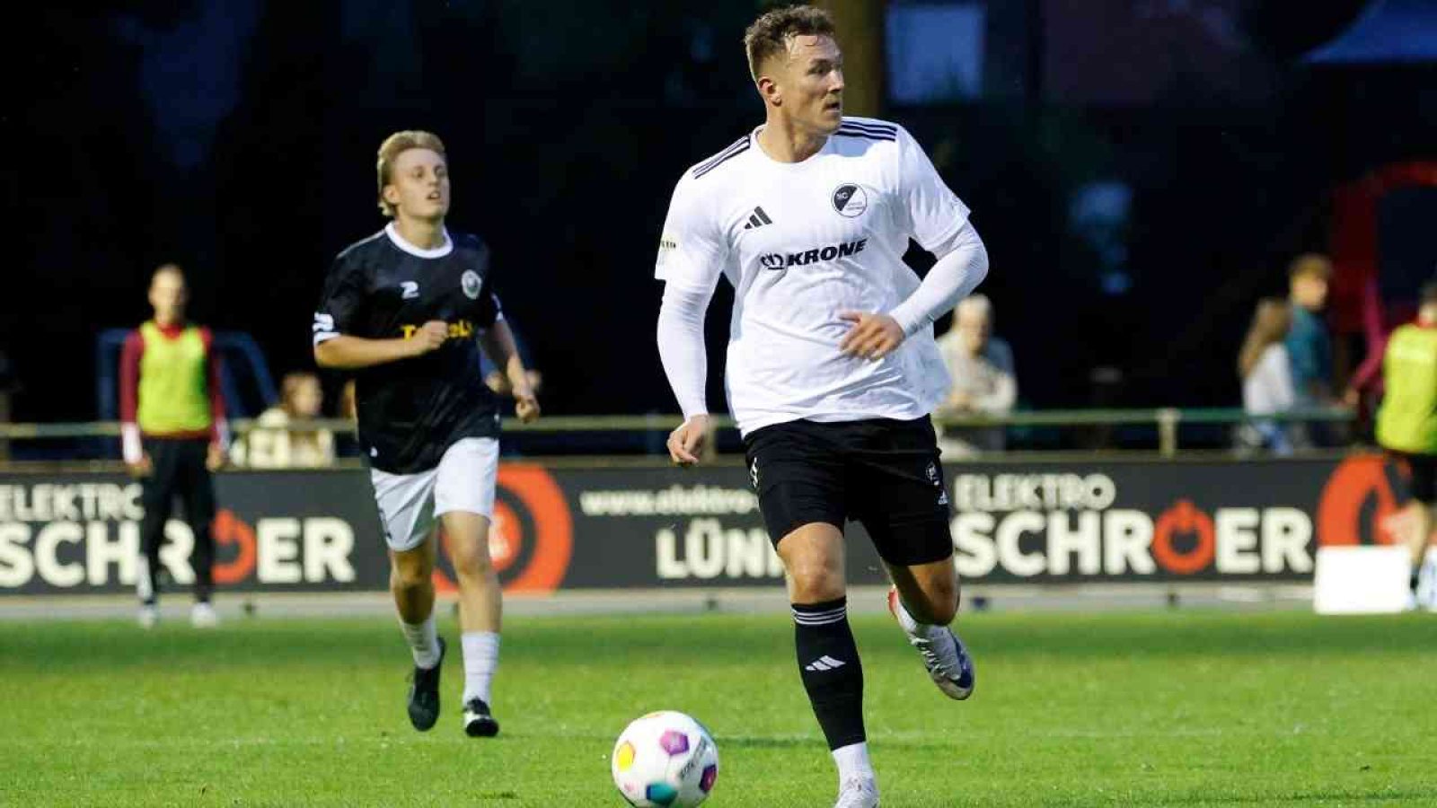 Ein Fußballspieler im weißen Trikot dribbelt den Ball auf dem Spielfeld, während ein Gegner im schwarzen Trikot verfolgt.