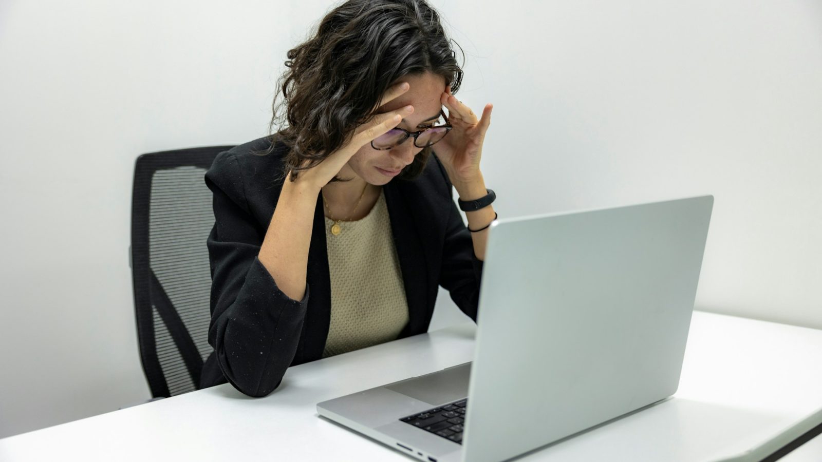 Erschöpfte Mitarbeiterin fasst sich mit schmerzverzerrtem Gesicht an den Kopf, im Hintergrund ihr Laptop am Schreibtisch