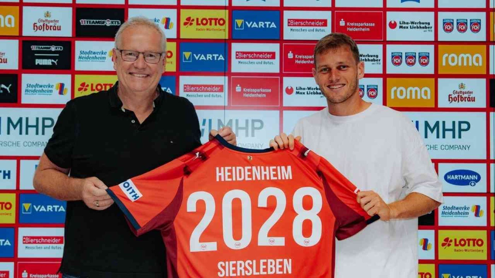 Zwei Männer halten ein Heidenheim-Trikot mit der Aufschrift 2028 und Siersleben vor einer Sponsorenwand.