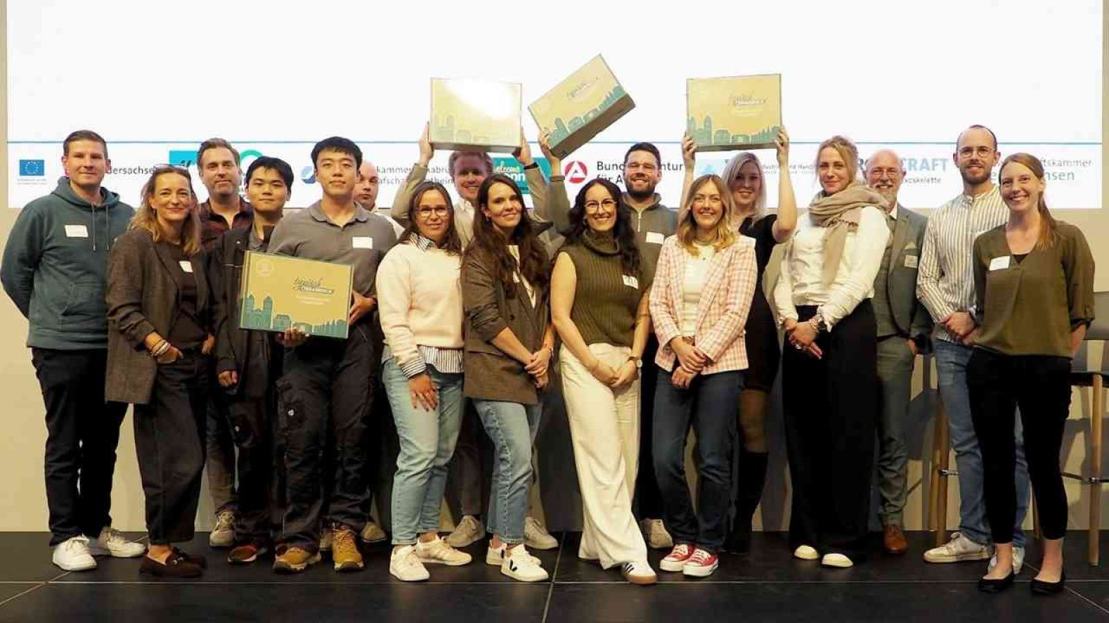 Gruppe von Menschen posiert bei einer Veranstaltung, einige halten Boxen hoch. Lächeln und Gruppenfoto auf Bühne.