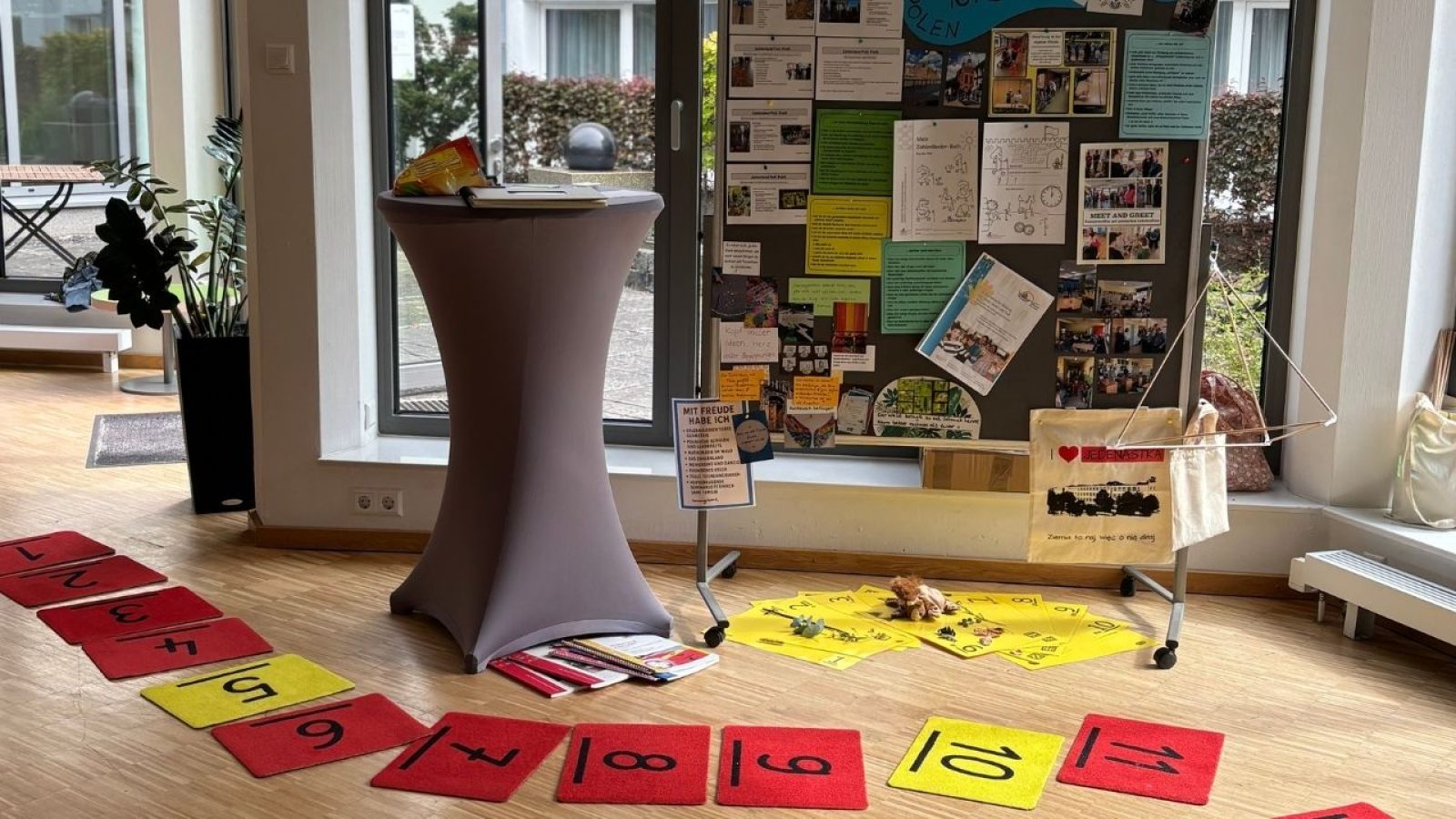 Informationsstand mit Pinnwand, Tisch und Zahlenmatten auf dem Boden in einem hellen Raum für Bildungsaktivitäten.