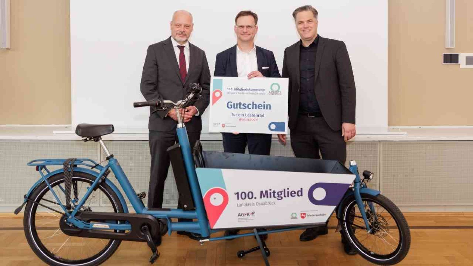 Drei Männer stehen neben einem blauen Lastenfahrrad und halten ein Schild mit der Aufschrift 'Gutschein für ein Lastenrad'.