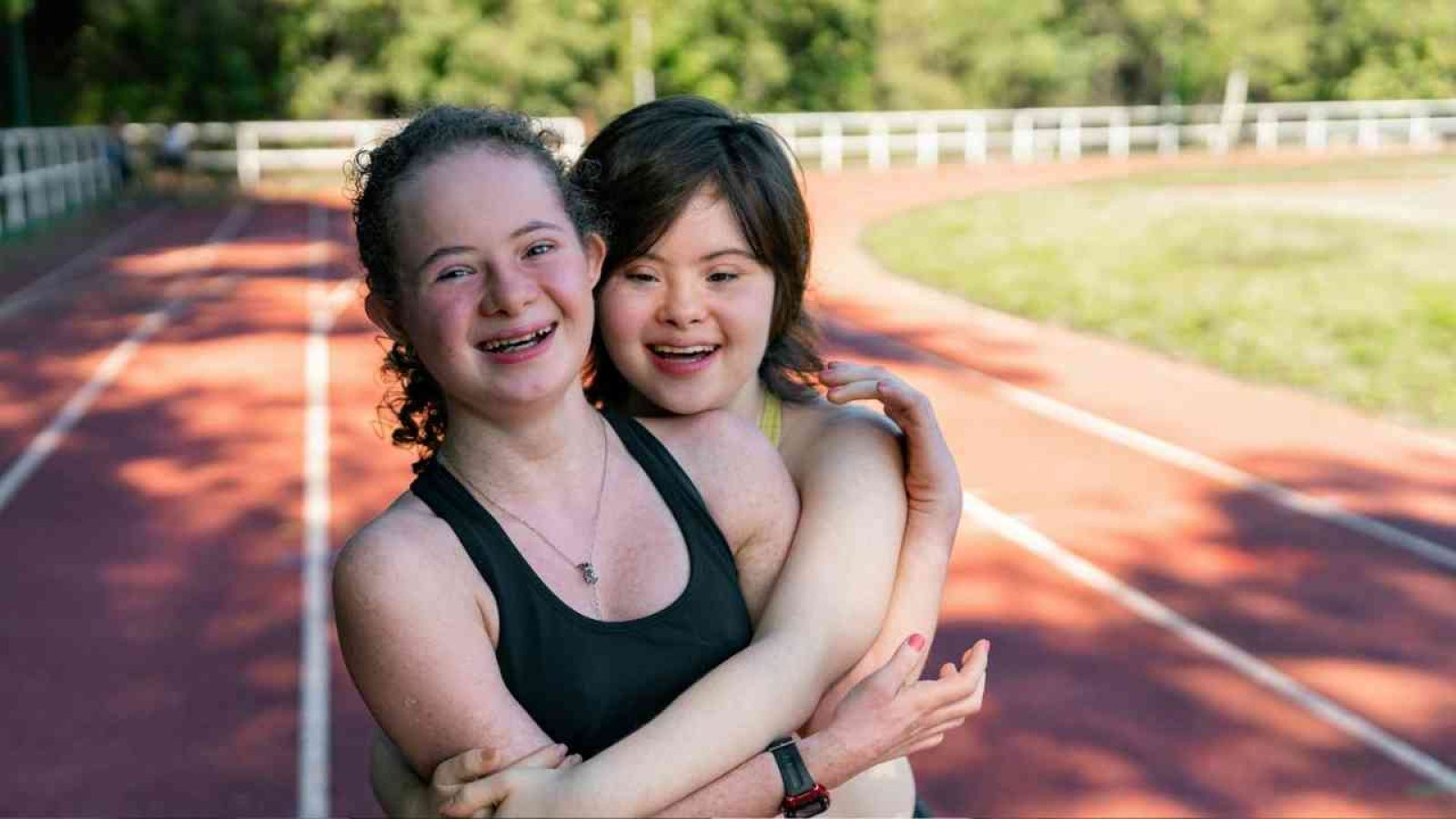 Zwei Mädchen mit Down-Syndrom umarmen sich auf einer Laufbahn im Freien.