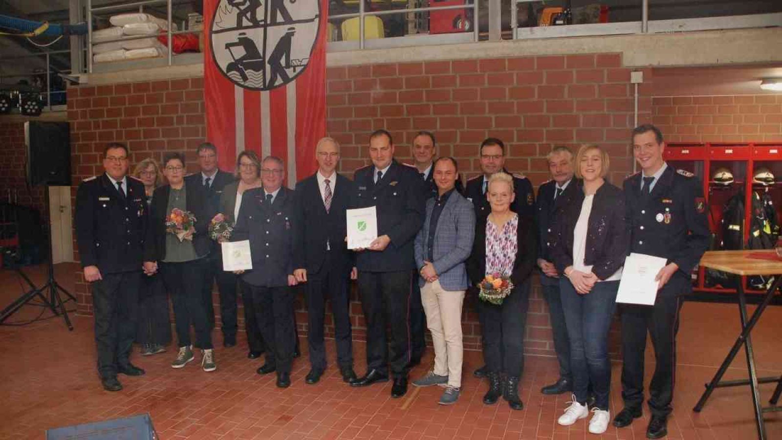 Gruppe von Feuerwehrleuten und Gästen bei einer Verabschiedung in einer Feuerwache mit Auszeichnungen und Blumen.