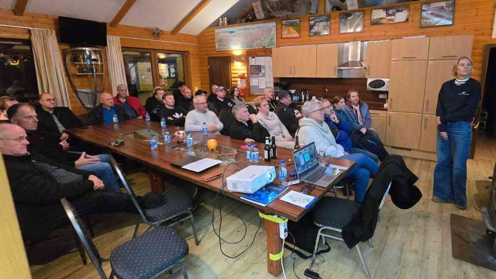 Gruppe von Personen, die in einem Raum an einem Vortrag oder einer Präsentation teilnehmen, mit einem Projektor und einem Tisch.