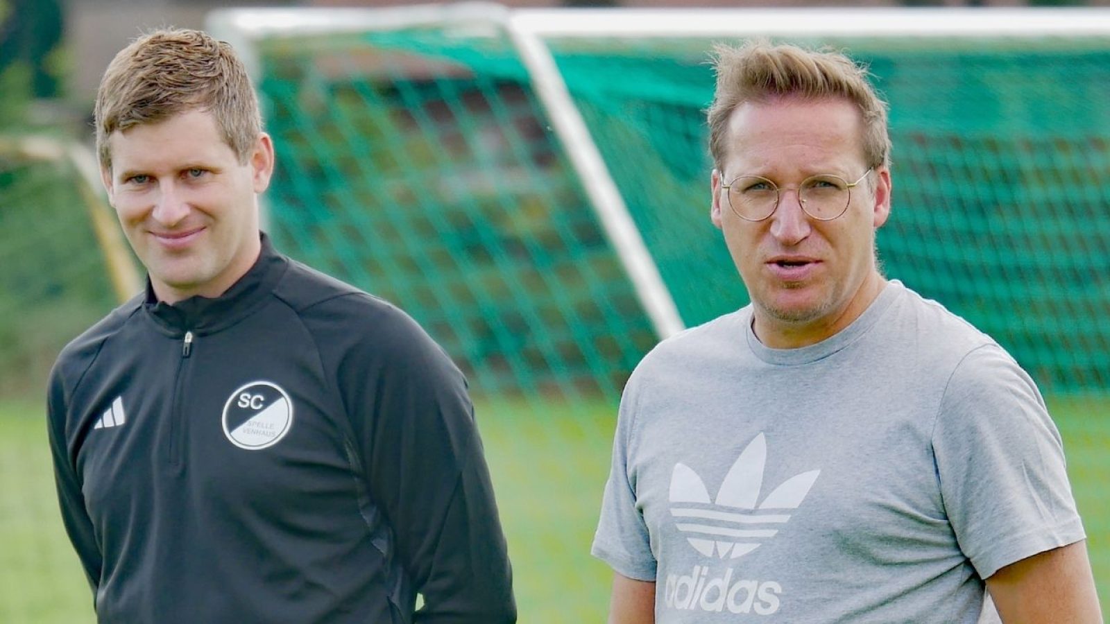 Zwei Männer vor einem Fußballtor im Freien, einer im Sportanzug, der andere im grauen Adidas T-Shirt.