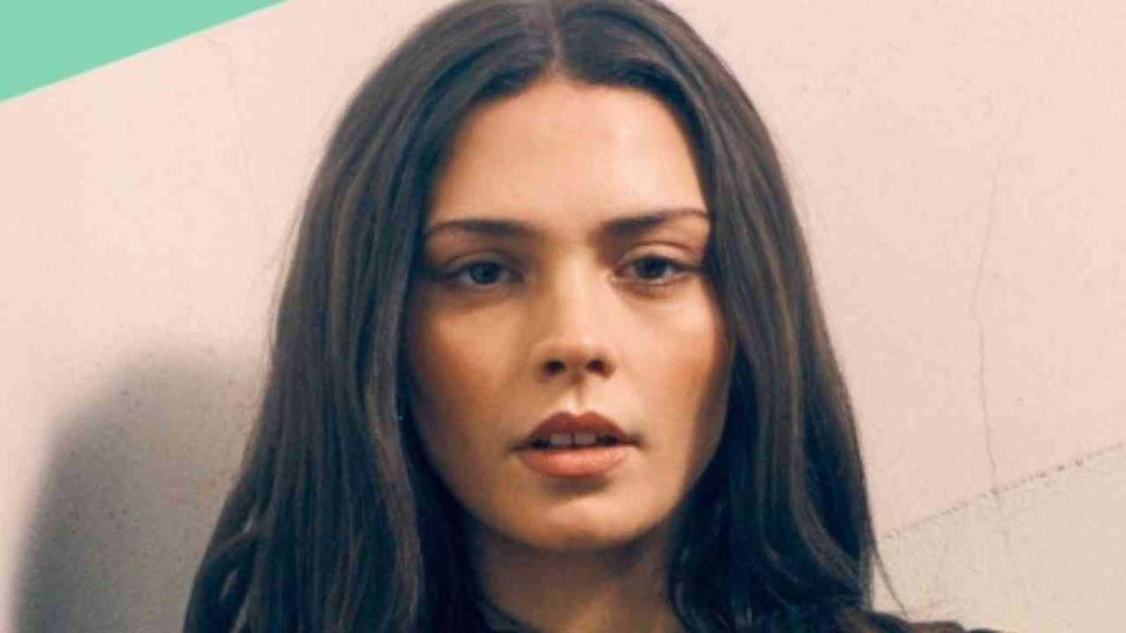 Porträt einer Frau mit langen, dunklen Haaren und neutralem Gesichtsausdruck vor einer hellen Wand.