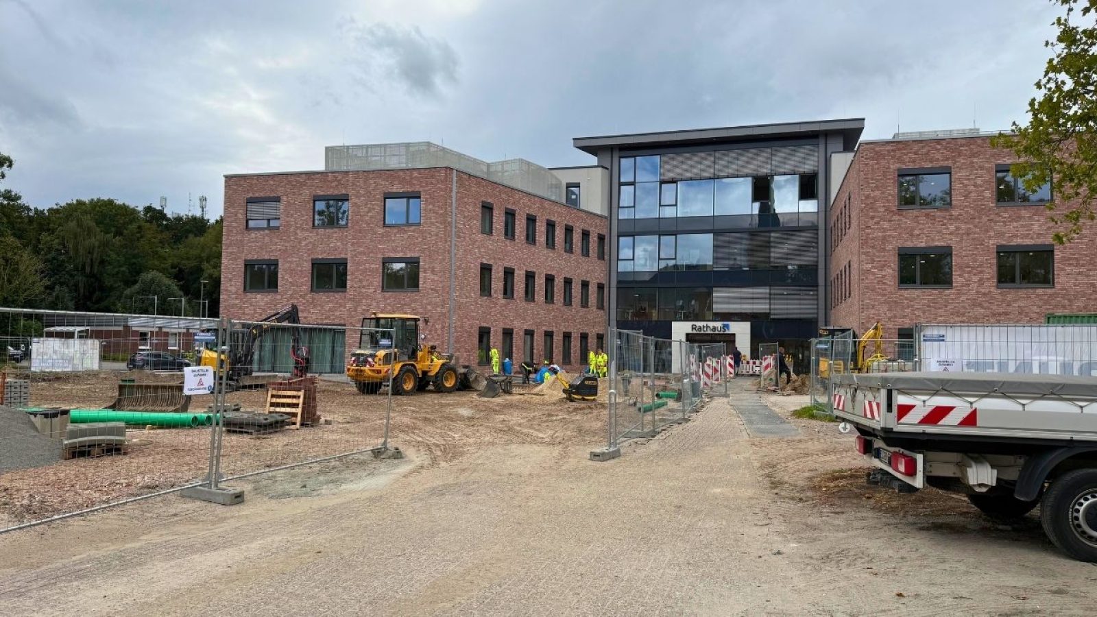 Baustelle vor modernem Gebäude in Ziegeloptik mit Bauarbeitern und Maschinen, umgeben von Bauzäunen.