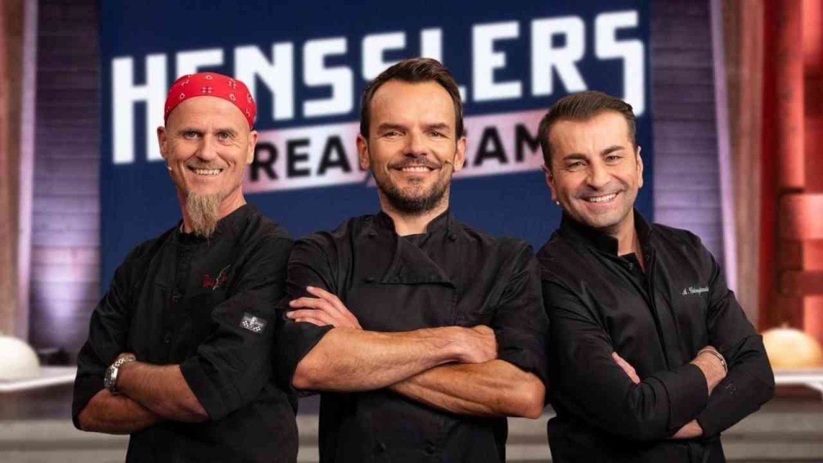 Drei Köche in schwarzen Jacken stehen lächelnd vor einem Hensslers Dream Team Hintergrund.