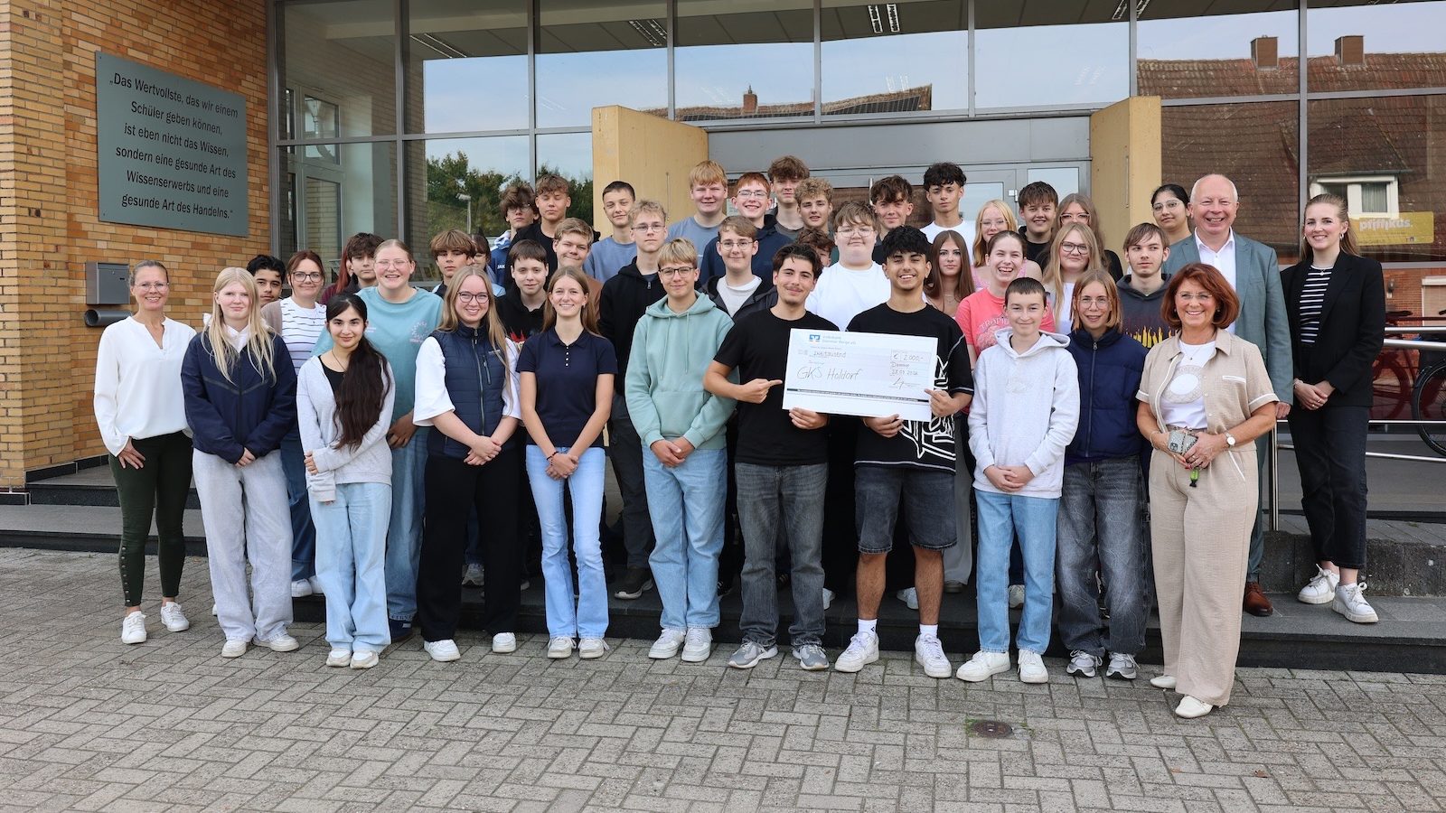 Schülerinnen und Schüler der Klassen 10a und 10b der Georg-Kerschensteiner-Schule Holdorf mit ihren Klassenlehrerinnen und Vertretern der Volksbank Dammer Berge vor dem Schulgebäude, in der Mitte ein Scheck über 2.000 Euro