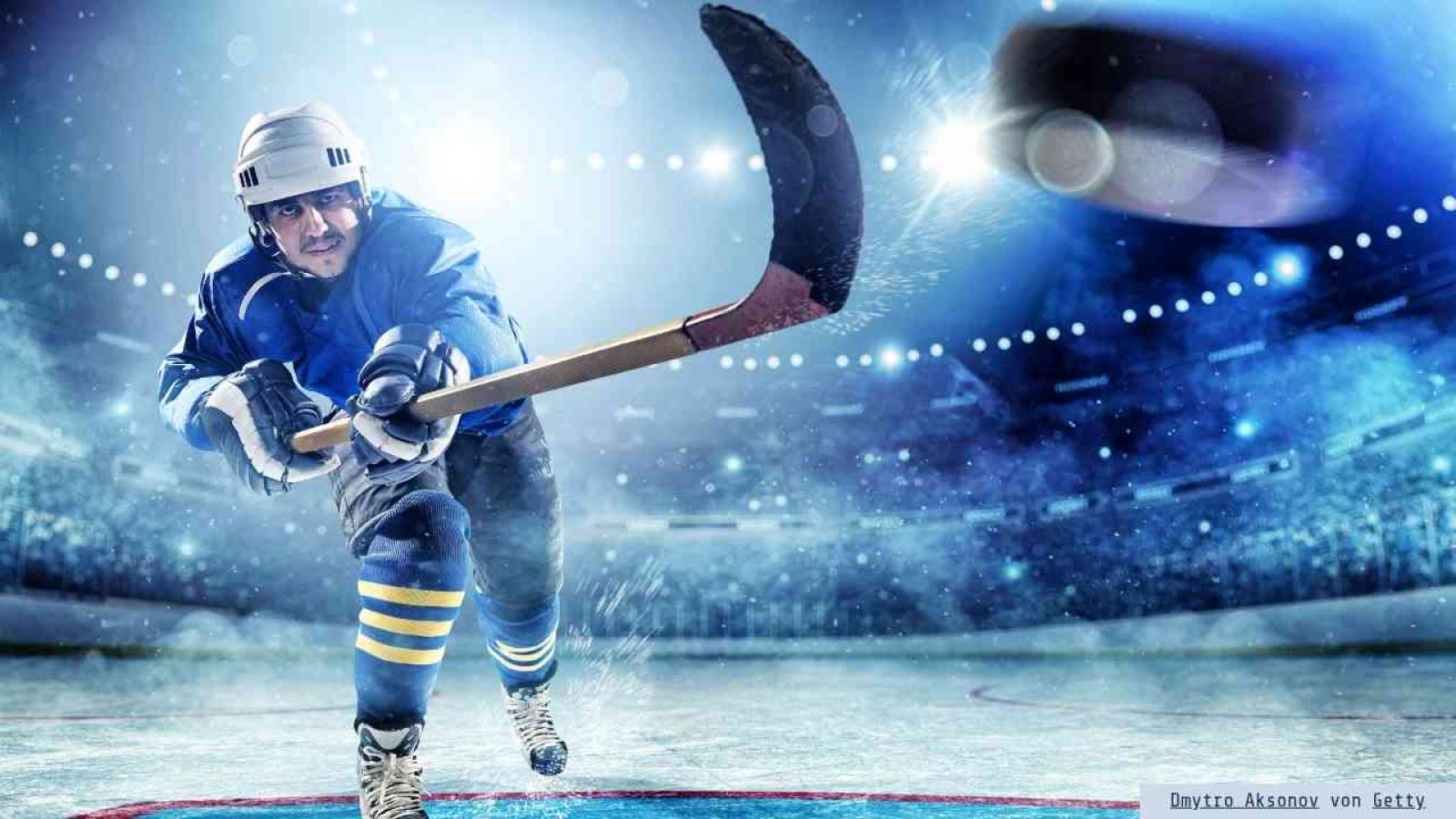 Eishockeyspieler in blauer Ausrüstung, der mit einem Schläger einen Puck auf dem Eis schießt.