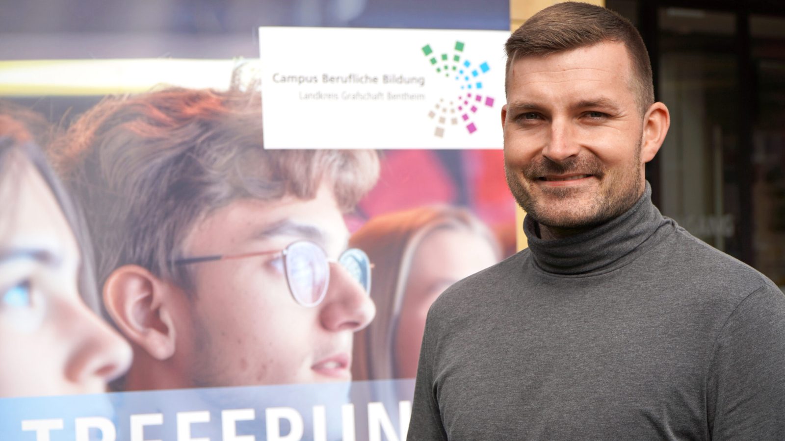 Mann lächelt vor Plakat des Campus Berufliche Bildung im Landkreis Grafschaft Bentheim, junge Menschen im Hintergrund.