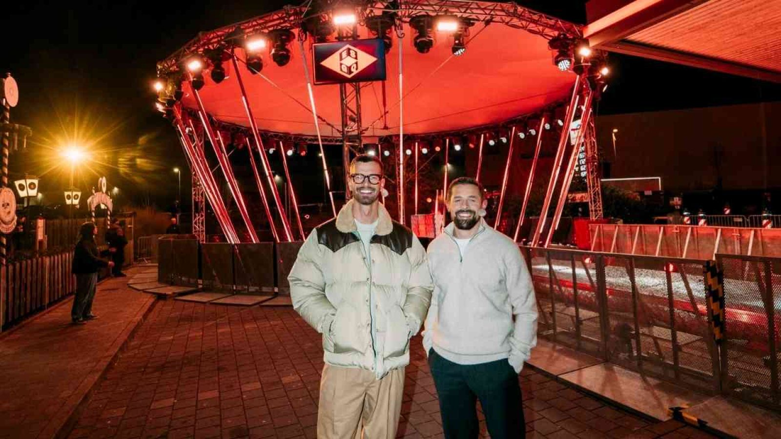 Zwei Männer bei Nacht vor einer rot beleuchteten Bühne auf einem Event im Freien, lächelnd in die Kamera.