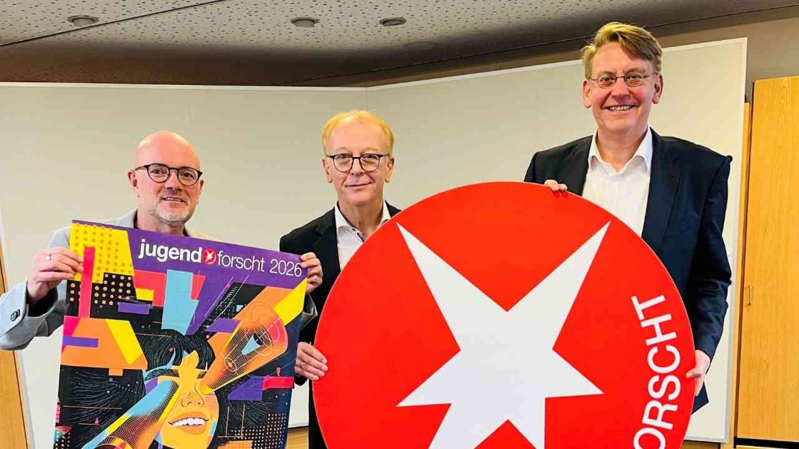 Drei Männer halten ein Plakat von 'Jugend forscht' und ein rotes Logo mit einem Stern in einem Besprechungsraum.