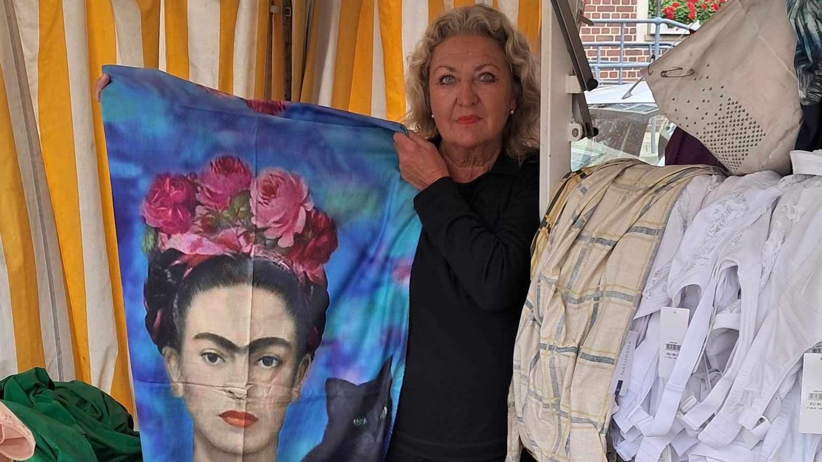 Händlerin Christa Endrigkeit hält an ihrem Marktstand ein bedrucktes Tuch hoch, davor Wäsche und Miederwaren