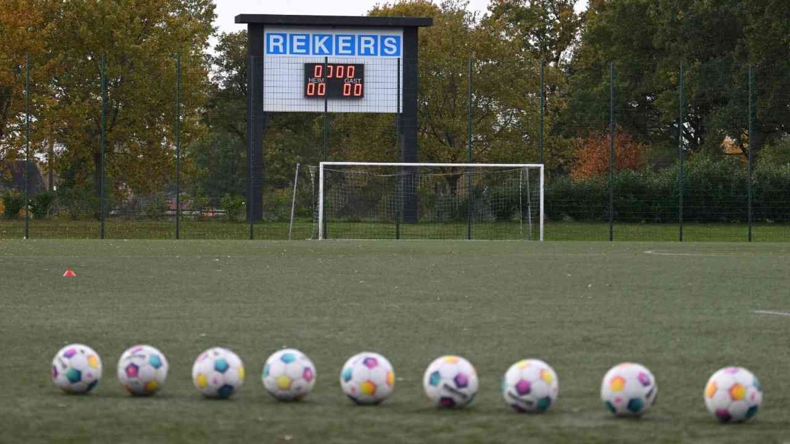Fußballfeld mit Tor und Anzeigetafel, die den Spielstand anzeigt, sowie einer Reihe von bunten Fußbällen im Vordergrund.