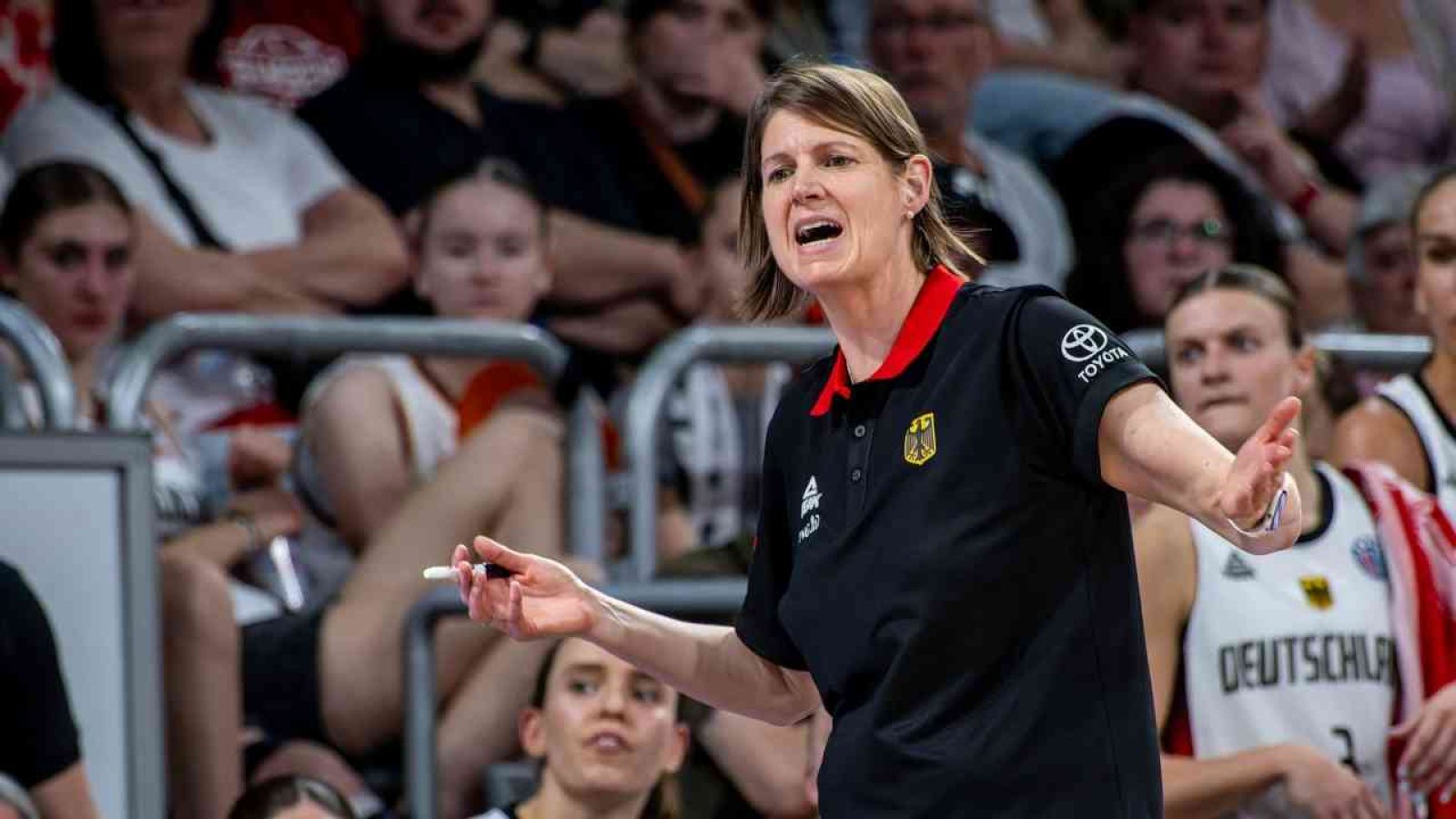 Basketballtrainerin mit einem schwarzen Polo und rotem Kragen, die während eines Spiels gestikuliert.