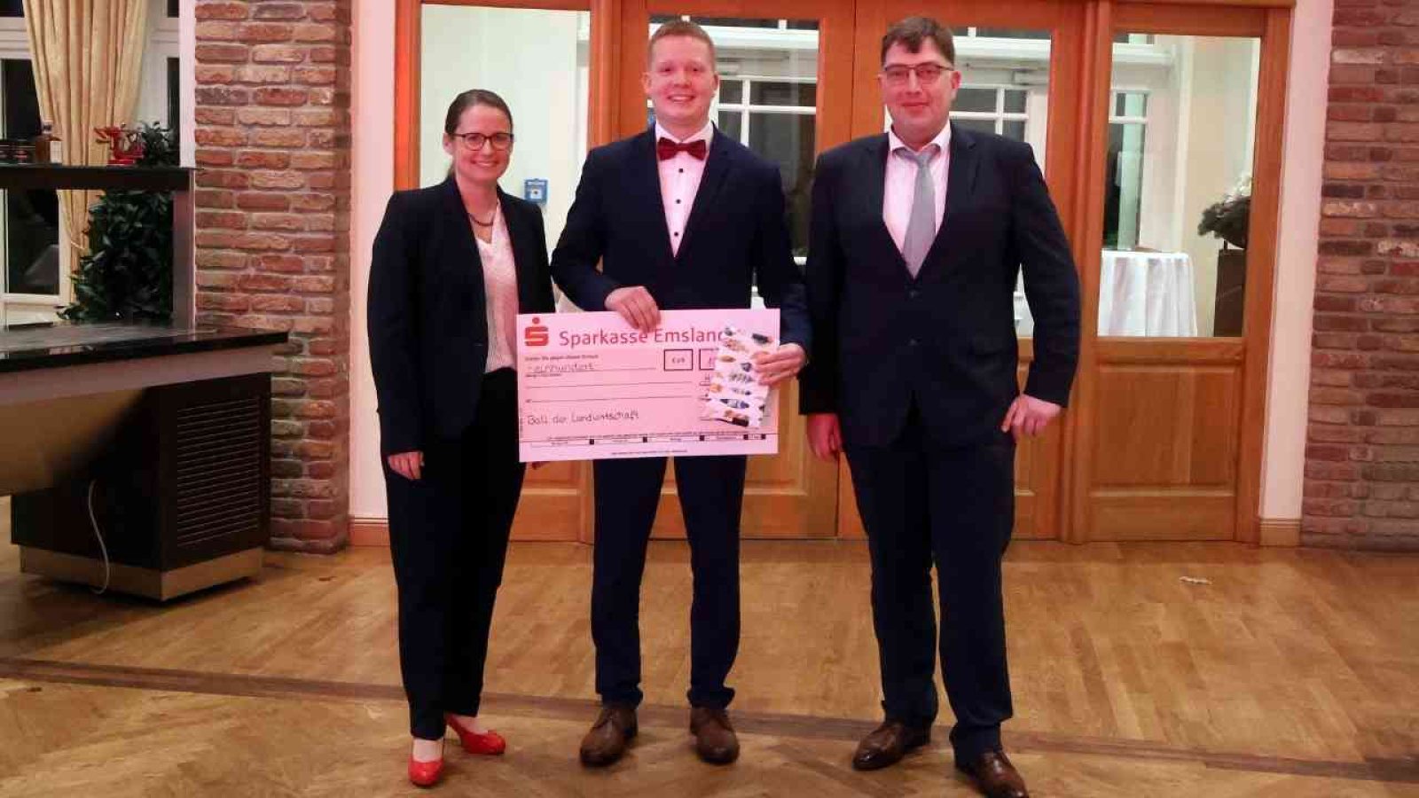 Drei Personen stehen in formeller Kleidung, während sie einen großen Scheck der Sparkasse Emsland präsentieren.