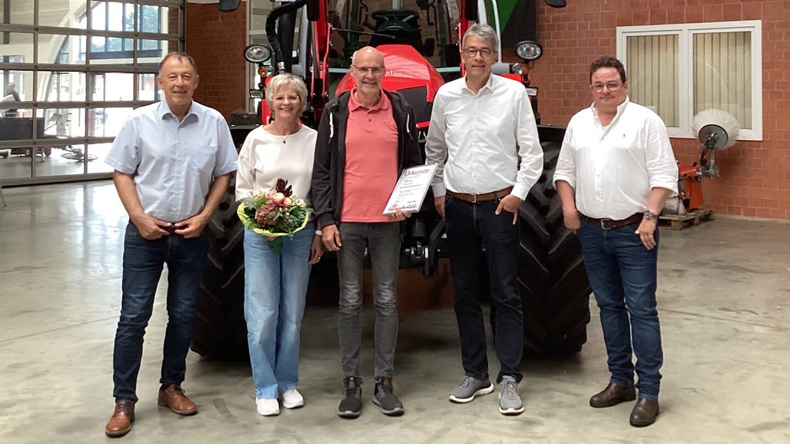 Jubilar Ludger Ahrens mit Urkunde, seine Frau Inge mit Blumenstrauß und drei Gratulanten vor einem roten Traktor in einer Werkhalle in Lastrup