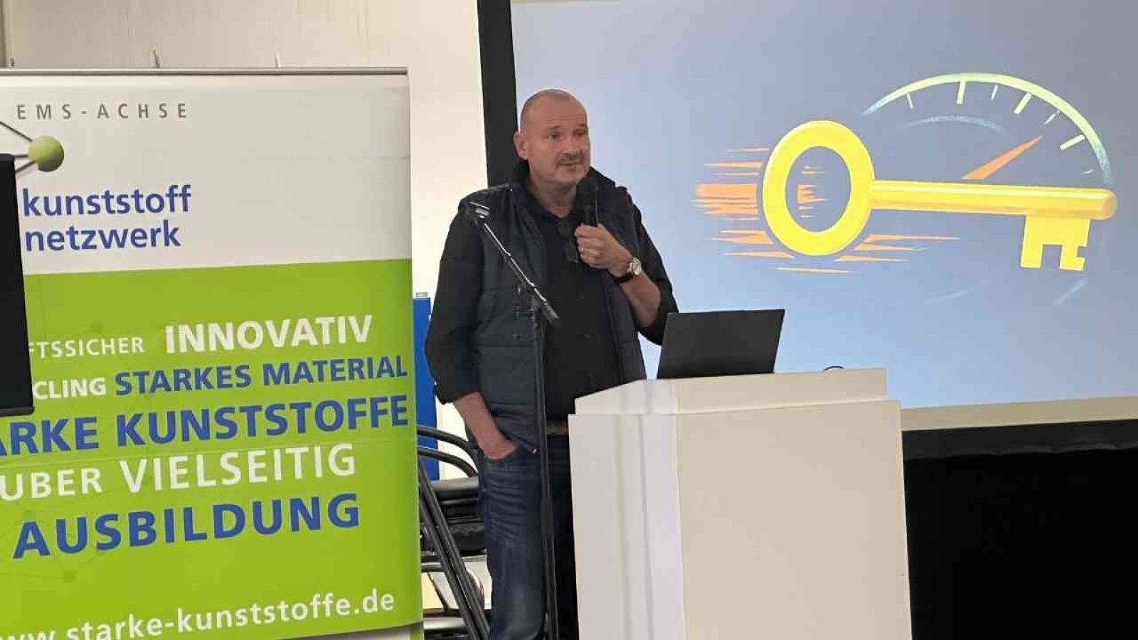 Mann spricht auf einer Veranstaltung mit einem Präsentationsbildschirm im Hintergrund, der einen Schlüssel und ein Geschwindigkeitsmesser zeigt.