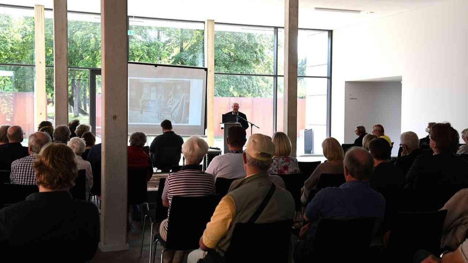Moderner Seminarraum mit Publikum und Vortragendem, der eine Präsentation hält. Blick auf Leinwand und große Fenster.