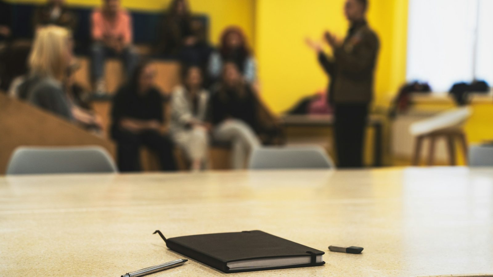 Notizblock und Stifte auf einem Tisch, im Hintergrund unscharf ein Redner und Publikum