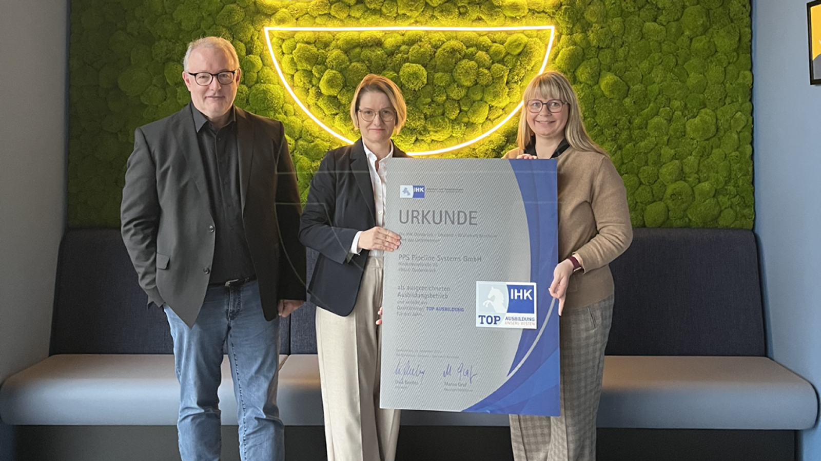 Drei Personen halten eine Urkunde der IHK in einem modernen Innenraum mit grüner Wanddekoration.
