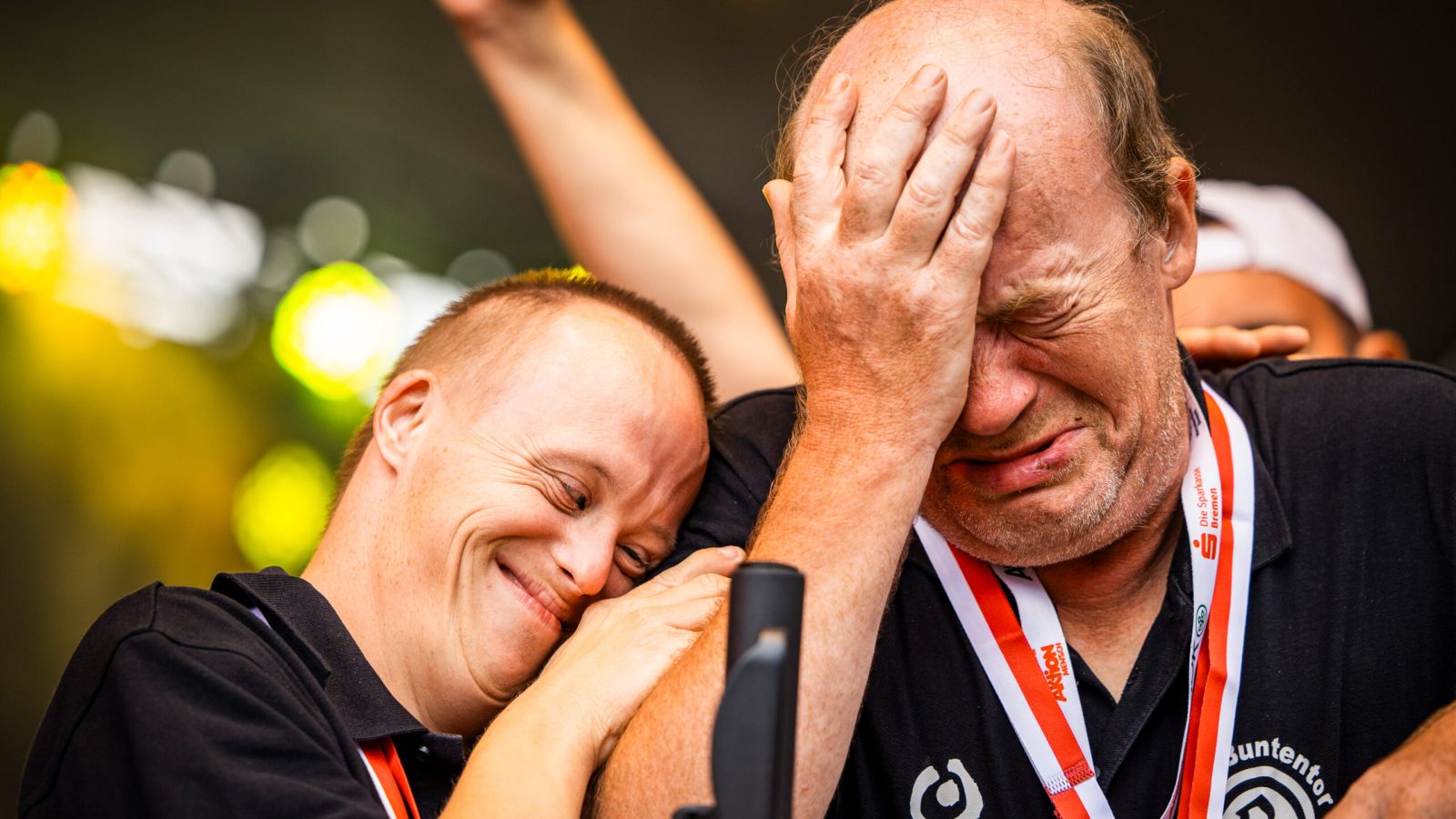 Zwei Männer mit Down-Syndrom, einer lächelt und der andere hält sich das Gesicht, beide tragen Medaillen.