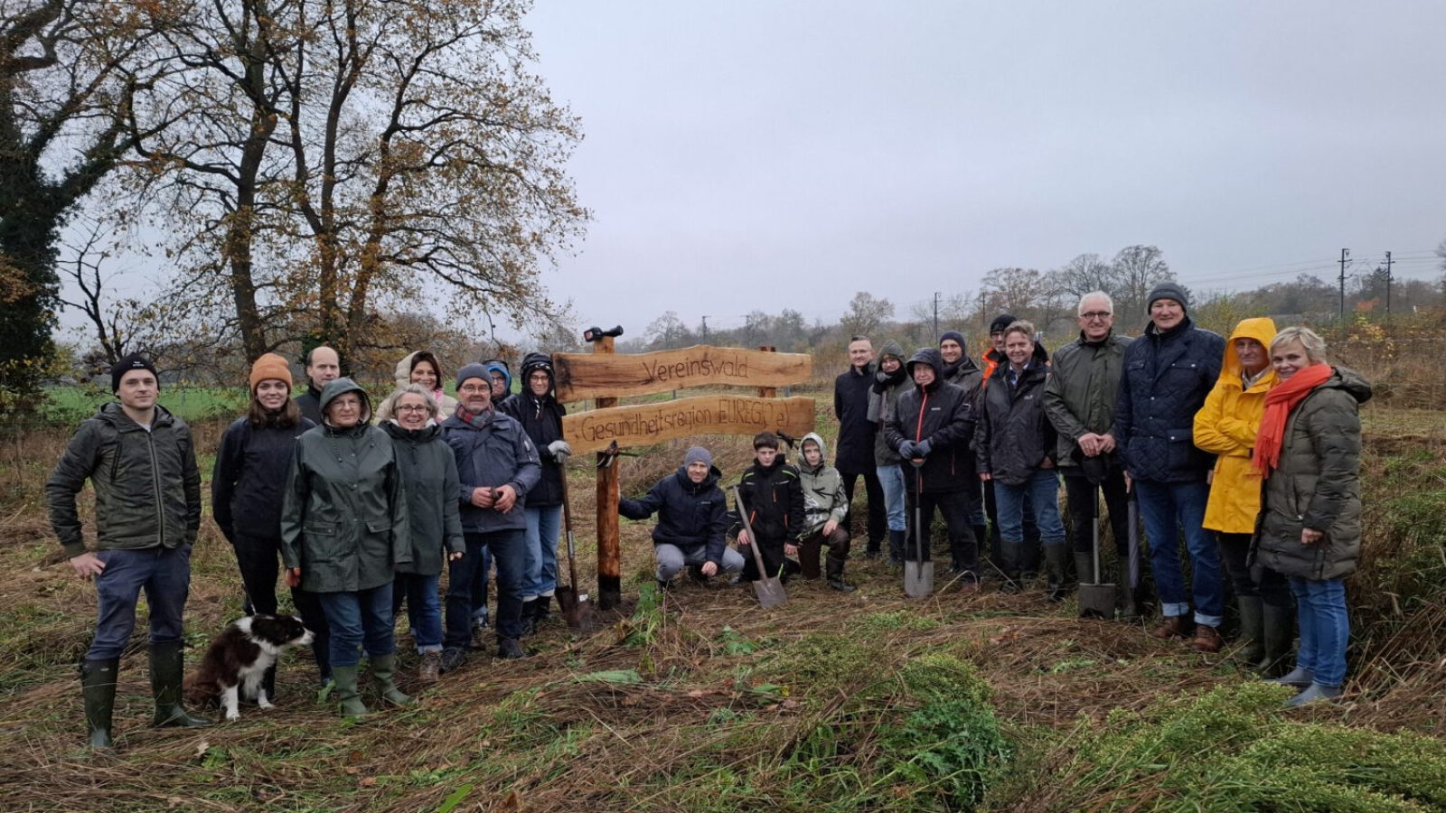 Gruppe von Menschen bei einer Pflanzaktion in der Gesundheitsregion Euregio mit Schaufeln und einem Holzschild im Hintergrund.