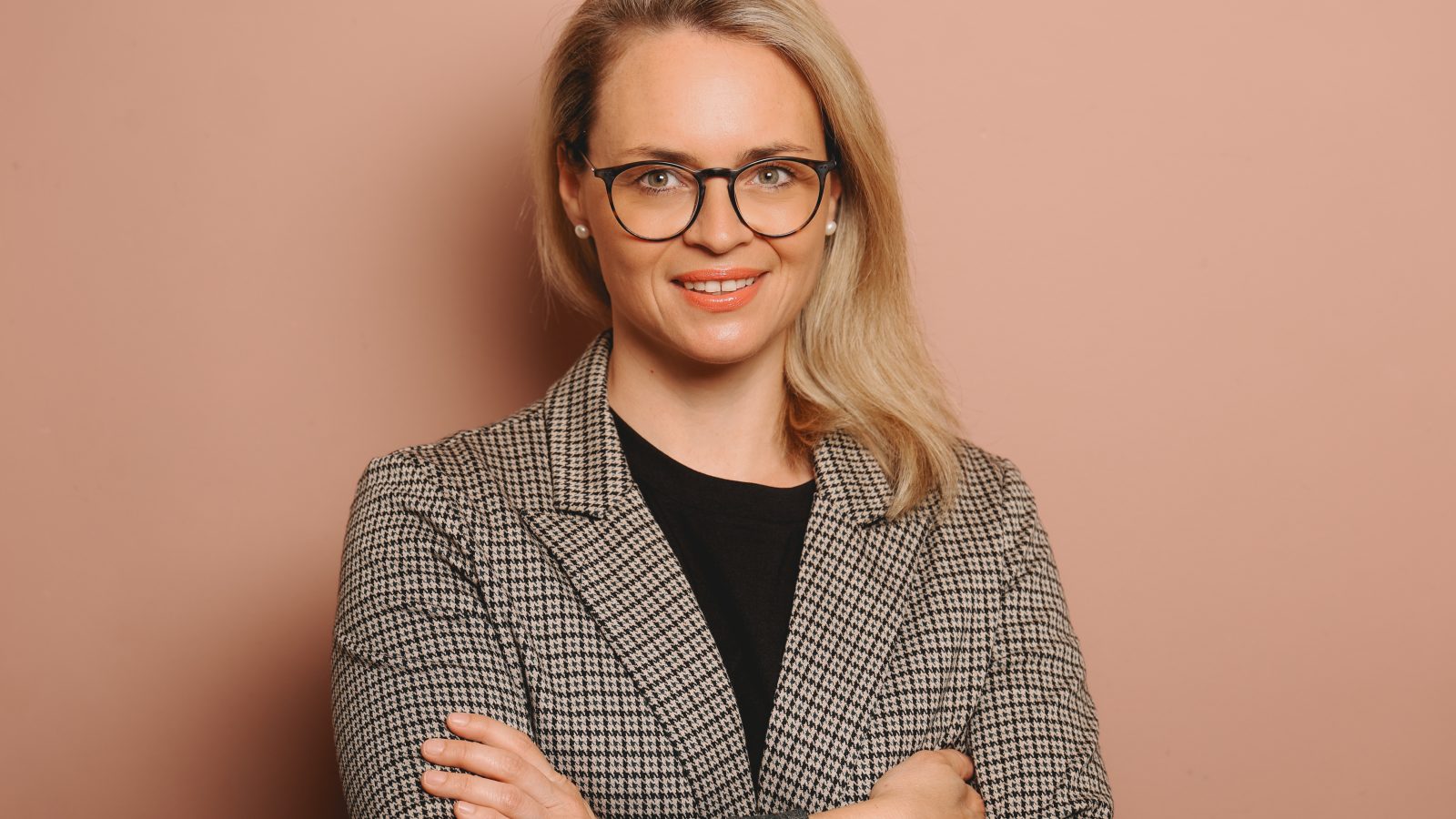 Porträt einer Frau mit blonden Haaren und Brille, die in einem karierten Blazer vor einem rosa Hintergrund steht.