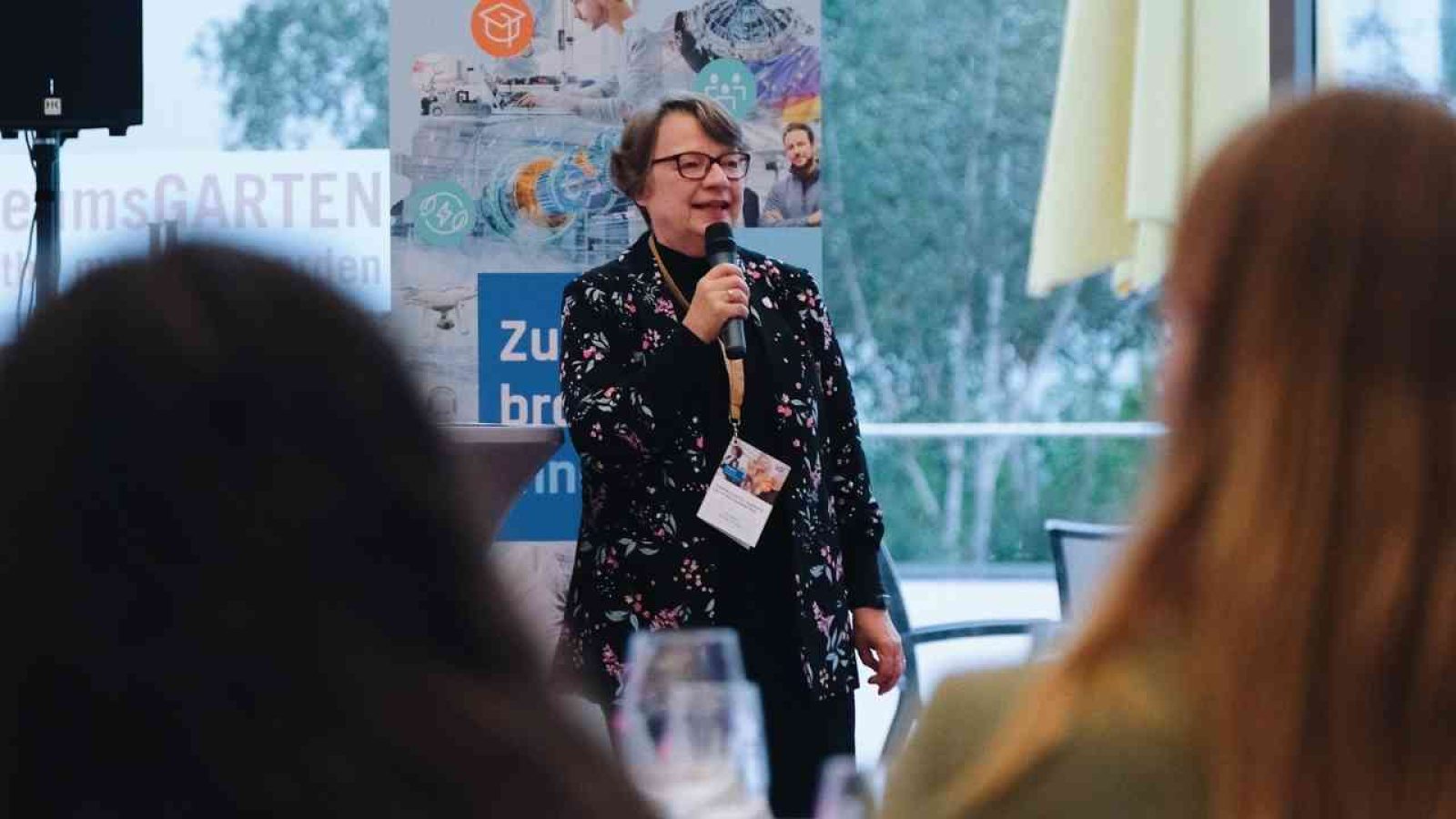 Person hält Vortrag auf Konferenz mit Mikrofon, vor Plakat; Publikum unscharf im Vordergrund.