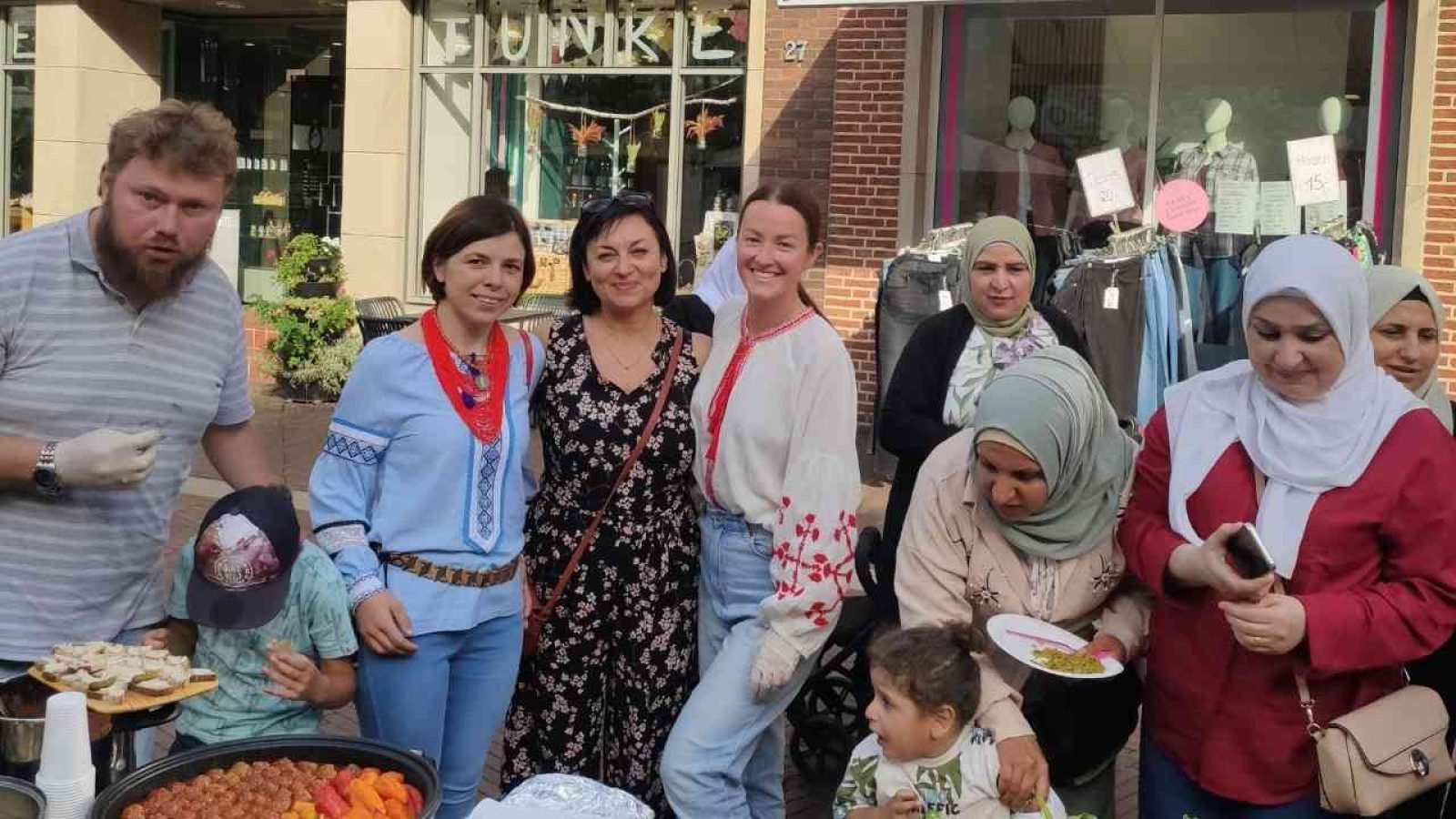 Gruppe von Menschen bei einem Straßenfest mit traditioneller Kleidung und Essen, fröhlich vereint vor einem Geschäft.