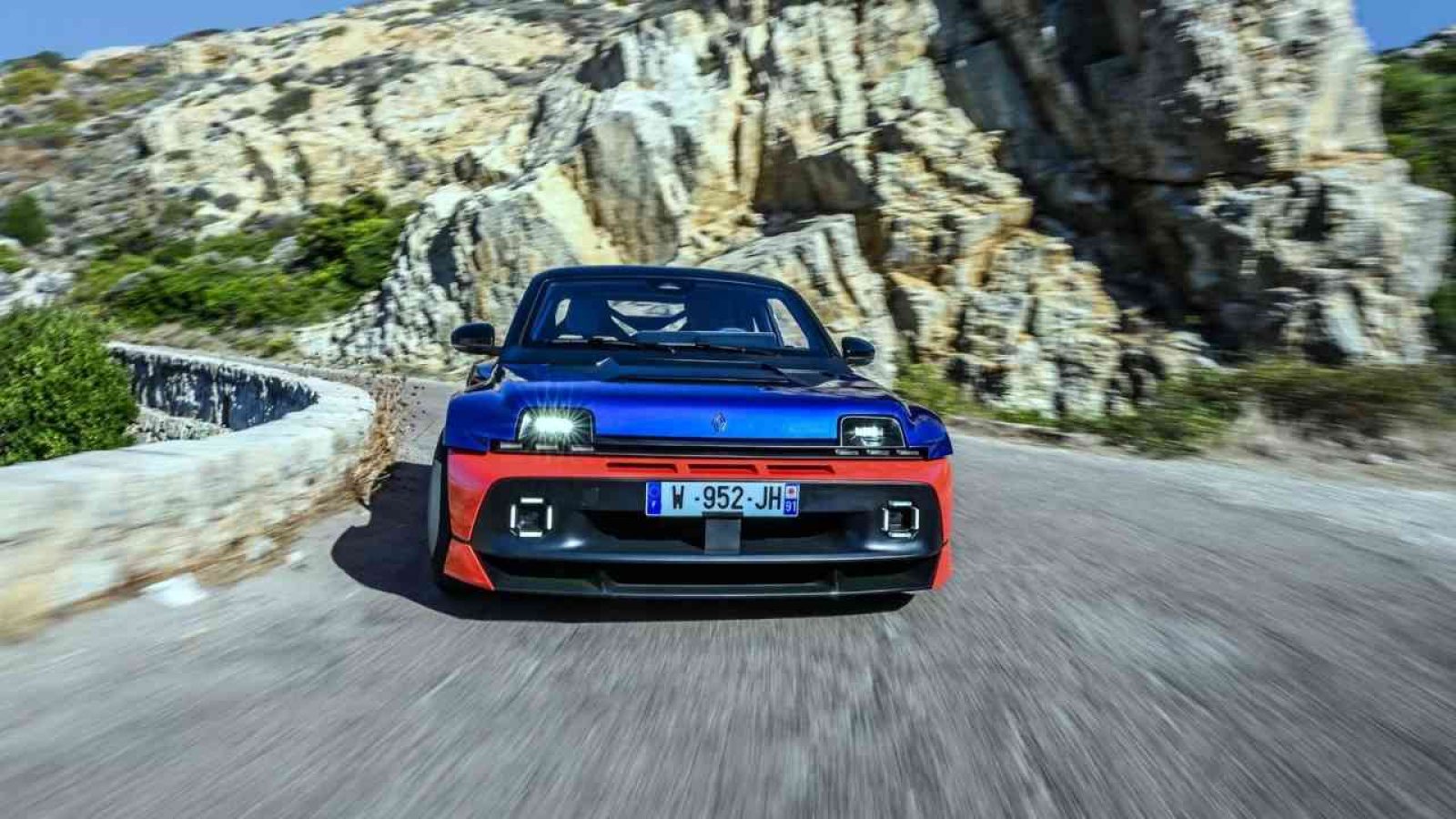 Renault 5 Turbo 3E in blauer und roter Lackierung auf einer kurvenreichen Straße.