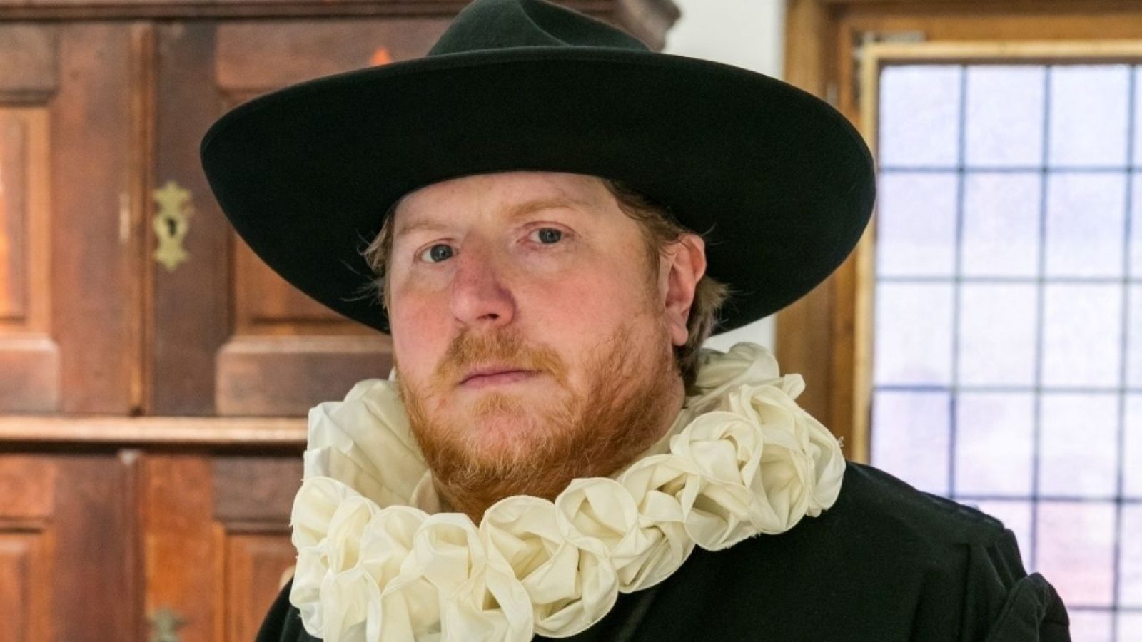 Mann mit einem schwarzen Hut und einem weißen Kragen, der in einem historischen Raum steht.