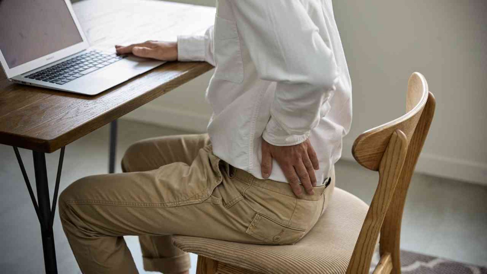 Person mit Rückenschmerzen beim Sitzen am Schreibtisch, Hand am unteren Rücken, Laptop auf Tisch.