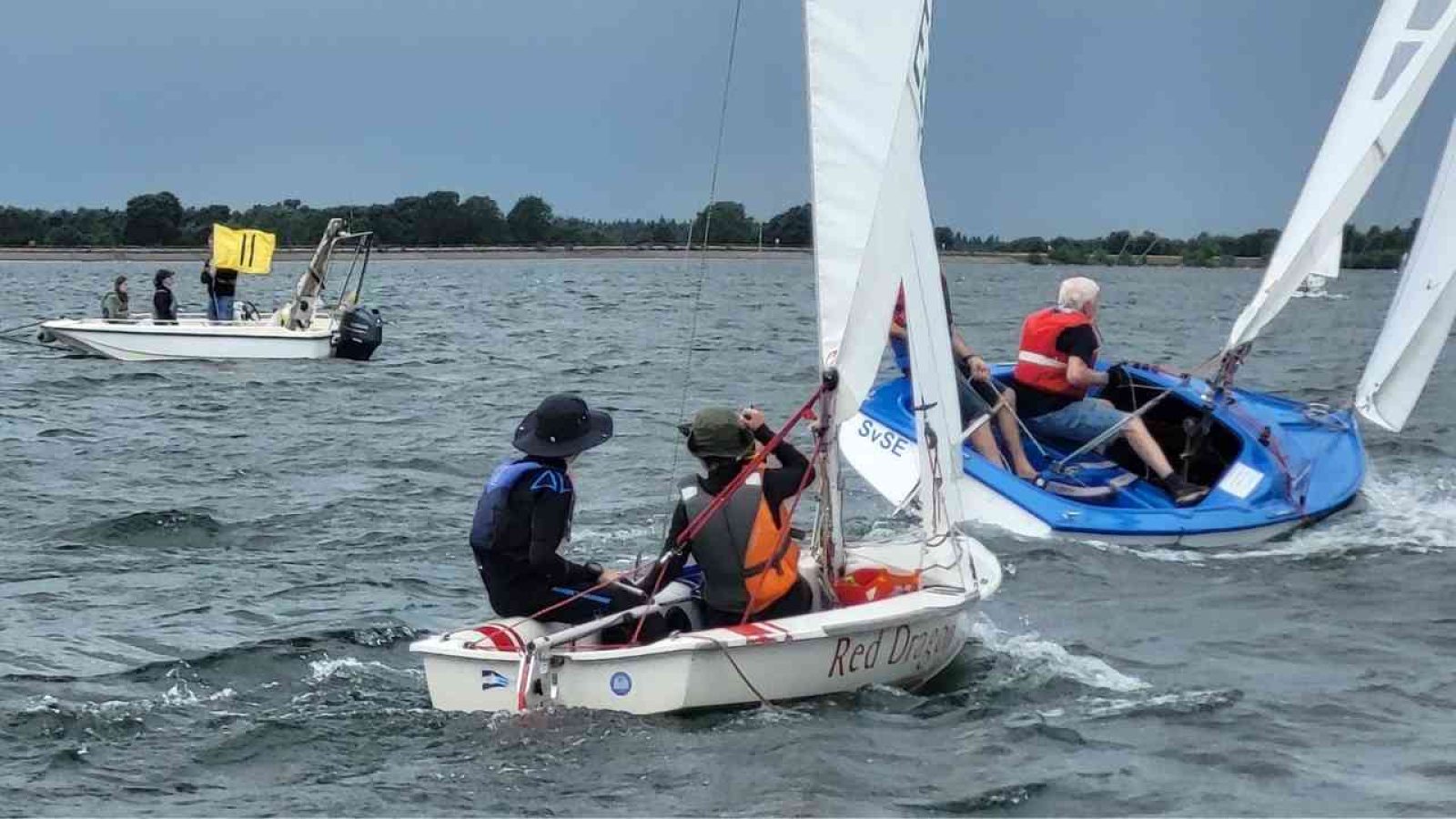 Segelboote bei Regatta auf dem Wasser, Personen in Schwimmwesten steuern Boote. Sicherheitsboot mit Crew in Nähe.