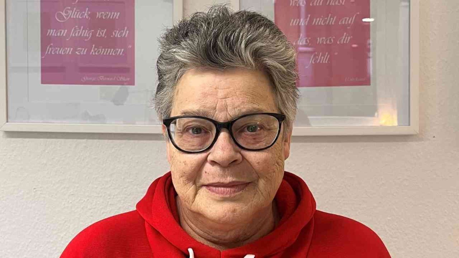 Ältere Frau mit Brille und grauen Haaren, die einen roten Pullover trägt, vor einer Wand mit gerahmten Bildern.
