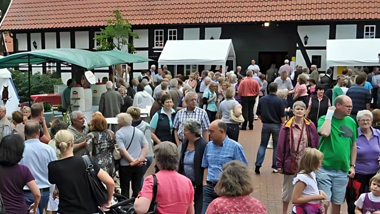 Menschenmenge bei einer Veranstaltung vor Fachwerkhaus, Verkaufsstände mit grünen und weißen Pavillons.