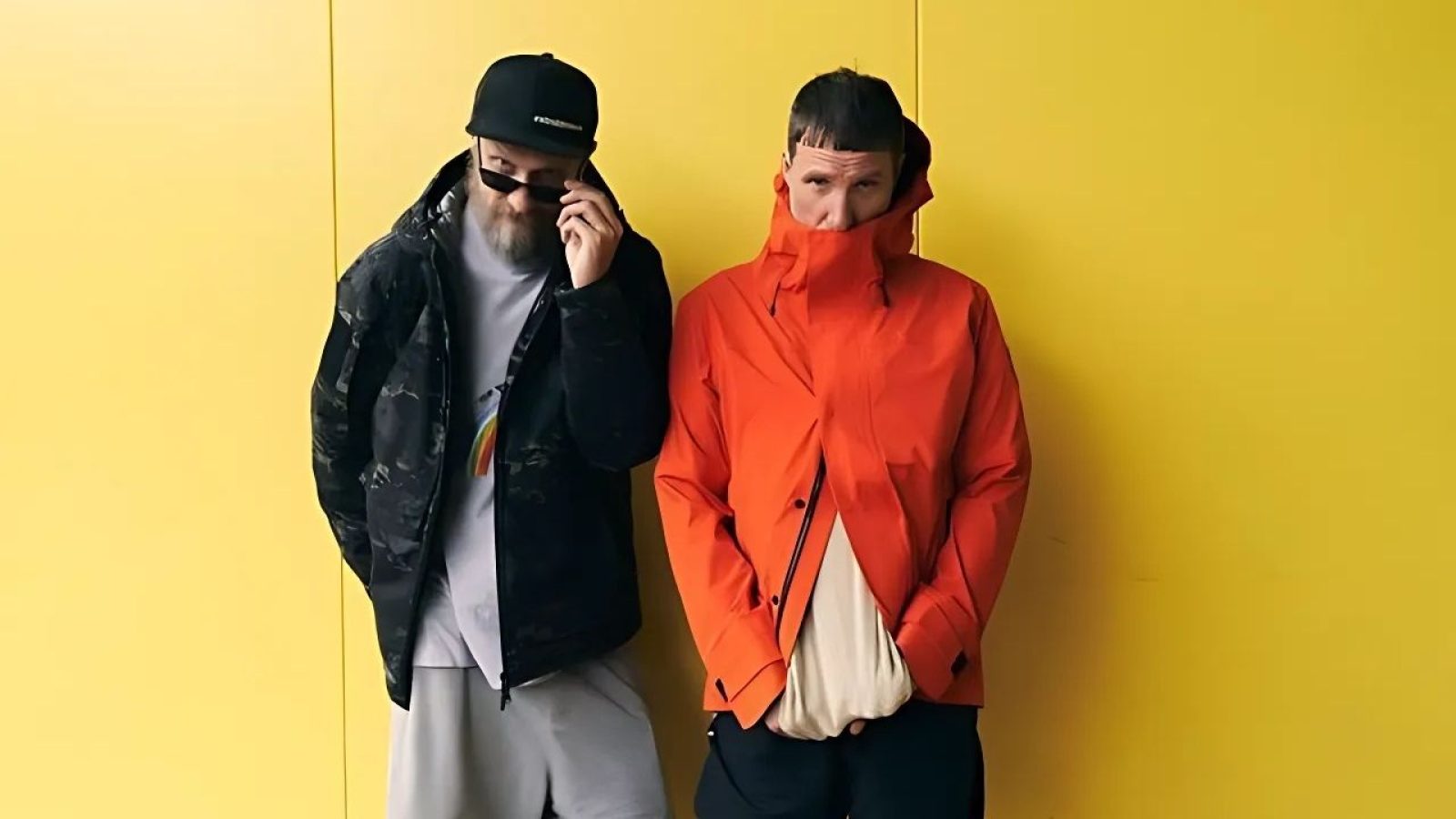 Zwei Männer in stylischen Jacken vor gelber Wand, links mit Sonnenbrille, rechts mit hochgezogenem Kragen.