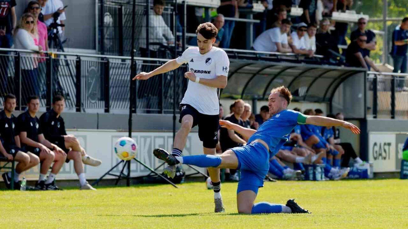 Ein Spieler in einem weißen Trikot tritt gegen einen Spieler in einem blauen Trikot während eines Fußballspiels.