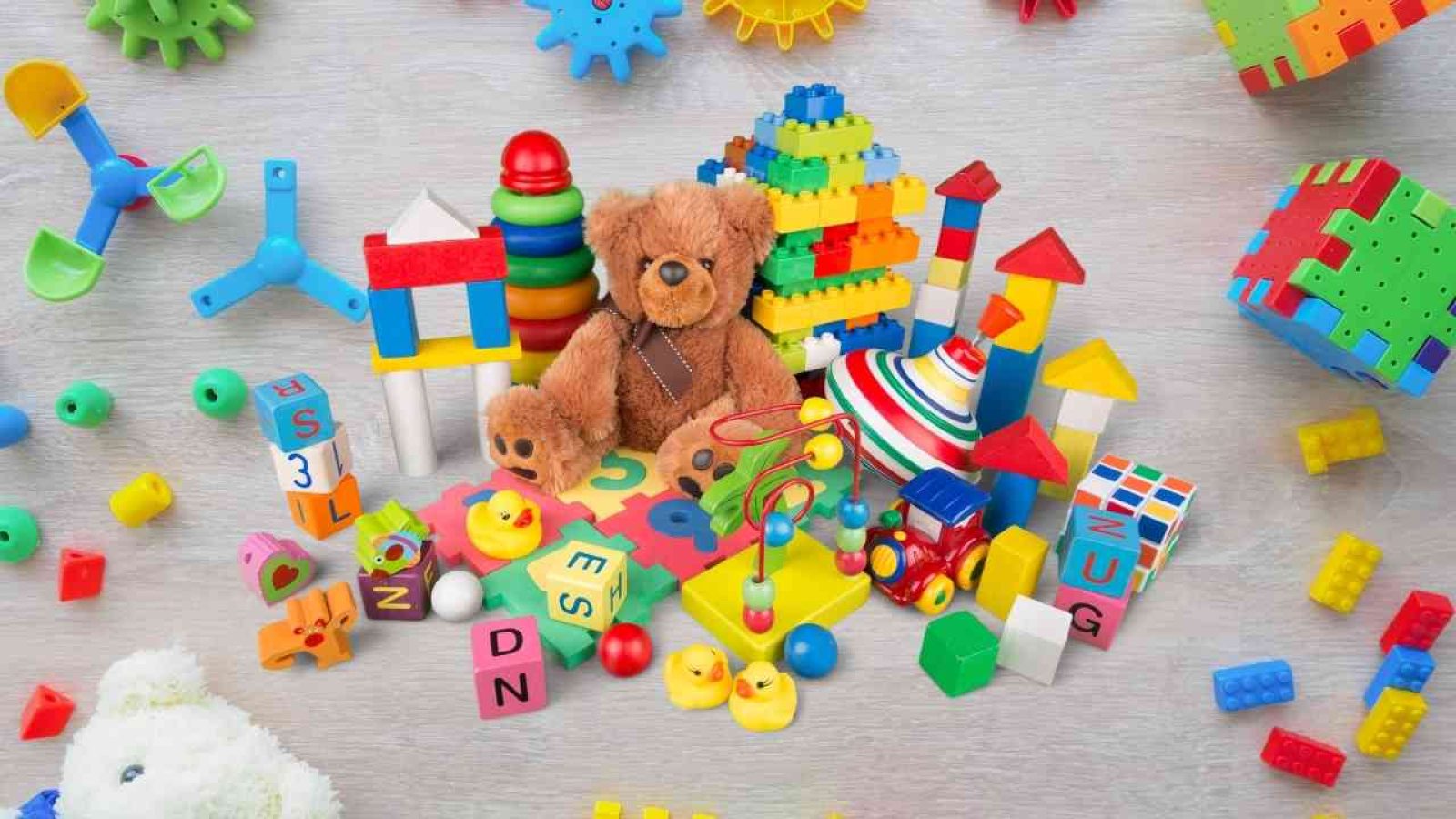 Bunte Kinderspielzeugsammlung mit Plüschteddy, Bauklötzen und Gummienten auf Holzfußboden.