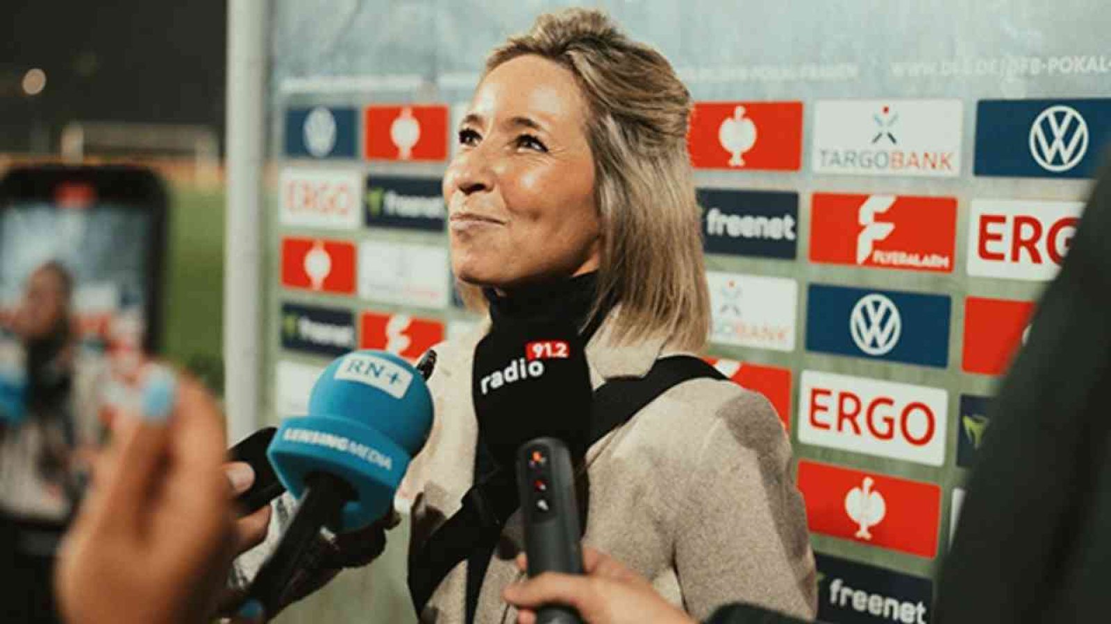 Dame bei Interview vor Sponsorenwand, hält Mikrophon mit blauem Logo, Stadionhintergrund unscharf.