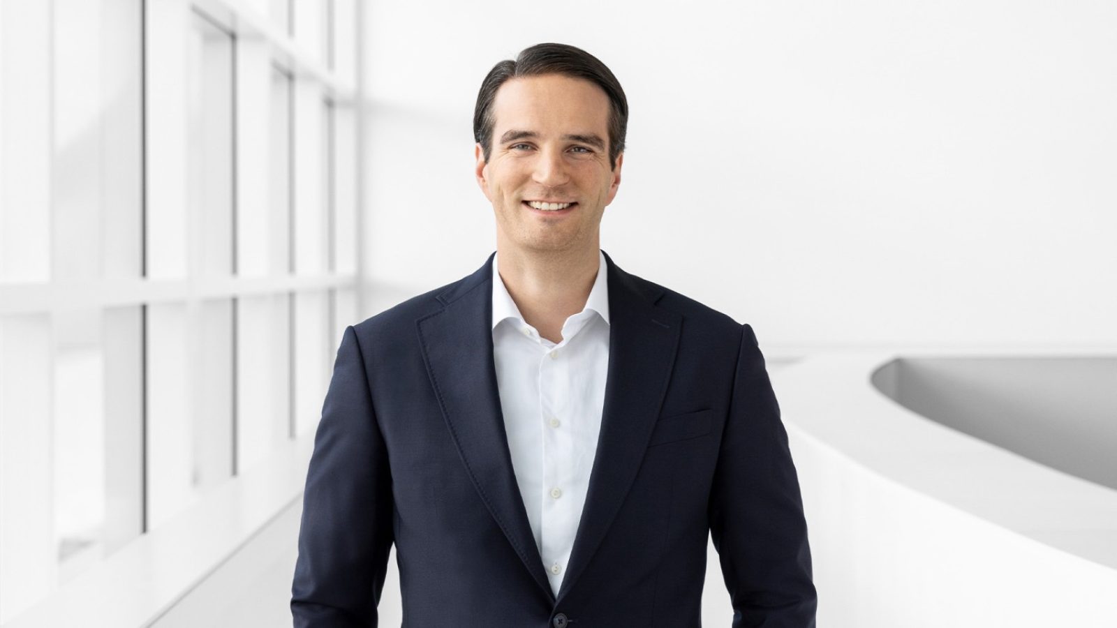 Mann in einem dunklen Anzug mit einem weißen Hemd, der in einem modernen Innenraum lächelt.