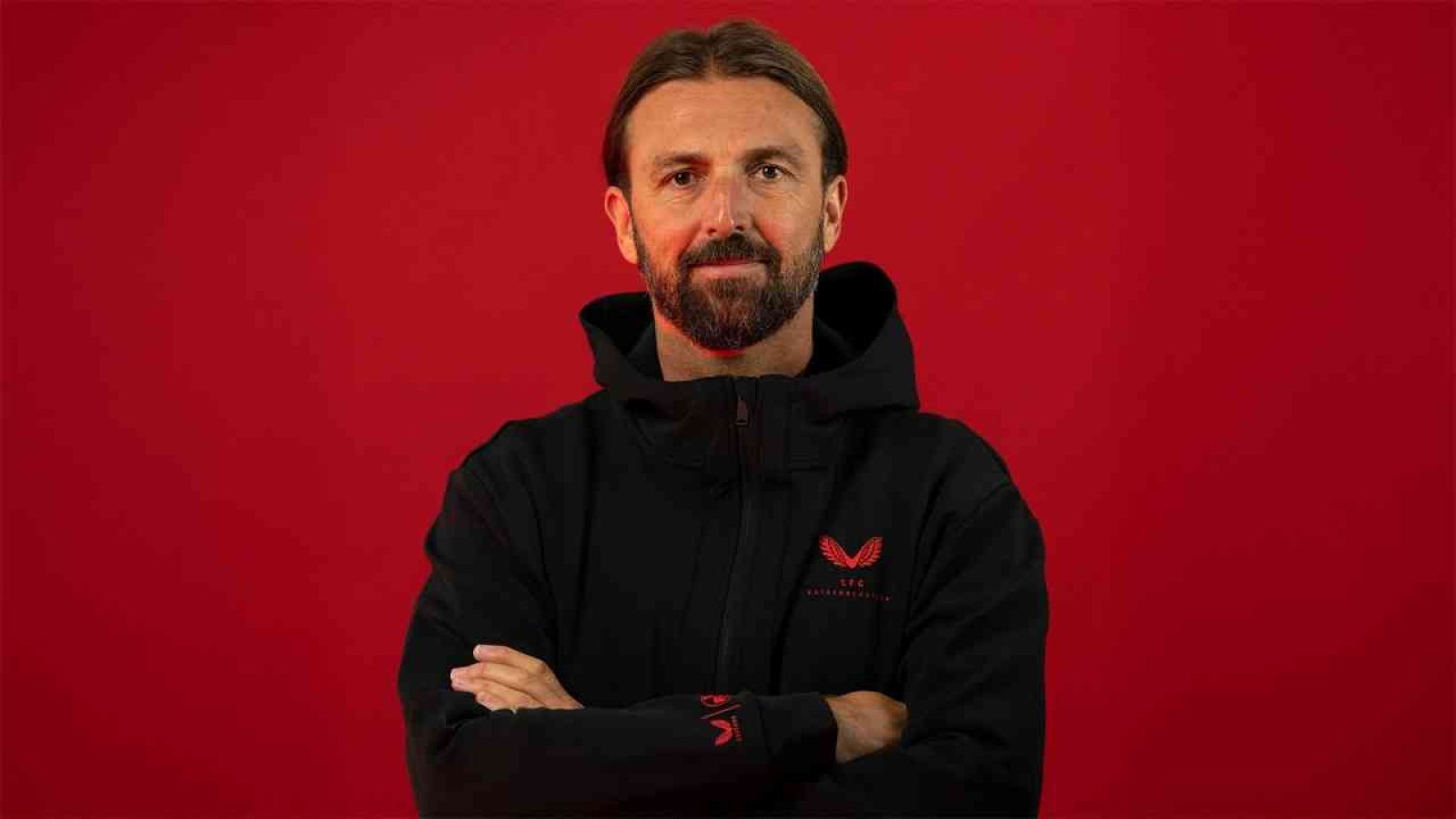 Mann mit Bart in schwarzer Jacke vor rotem Hintergrund, mit verschränkten Armen und neutralem Ausdruck.