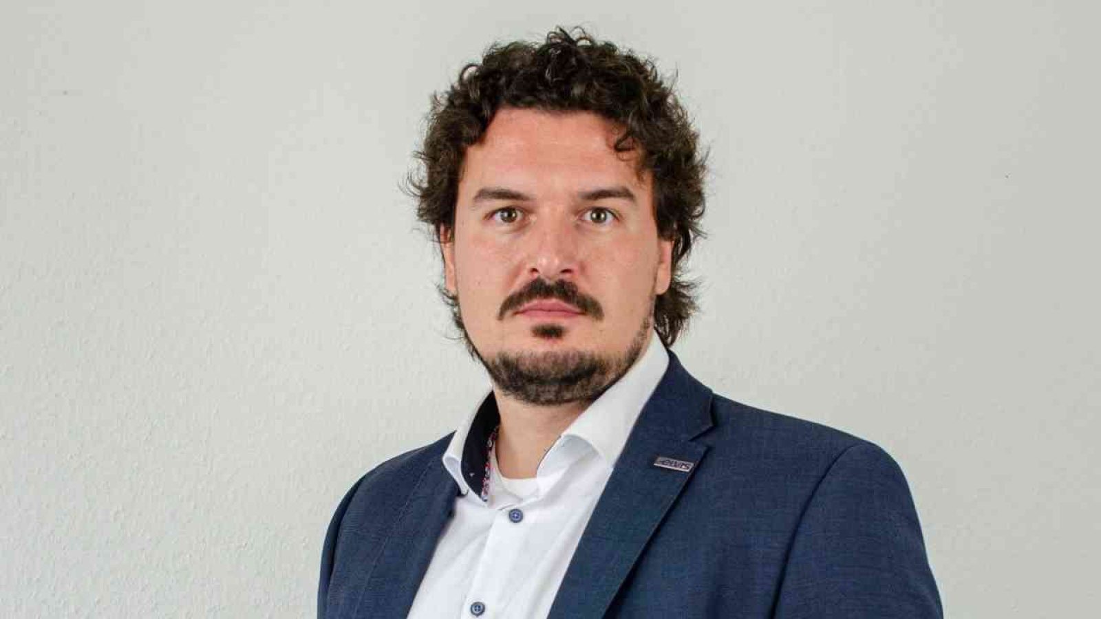 Mann mit lockigem Haar in einem Anzug, der direkt in die Kamera schaut, vor einem neutralen Hintergrund.