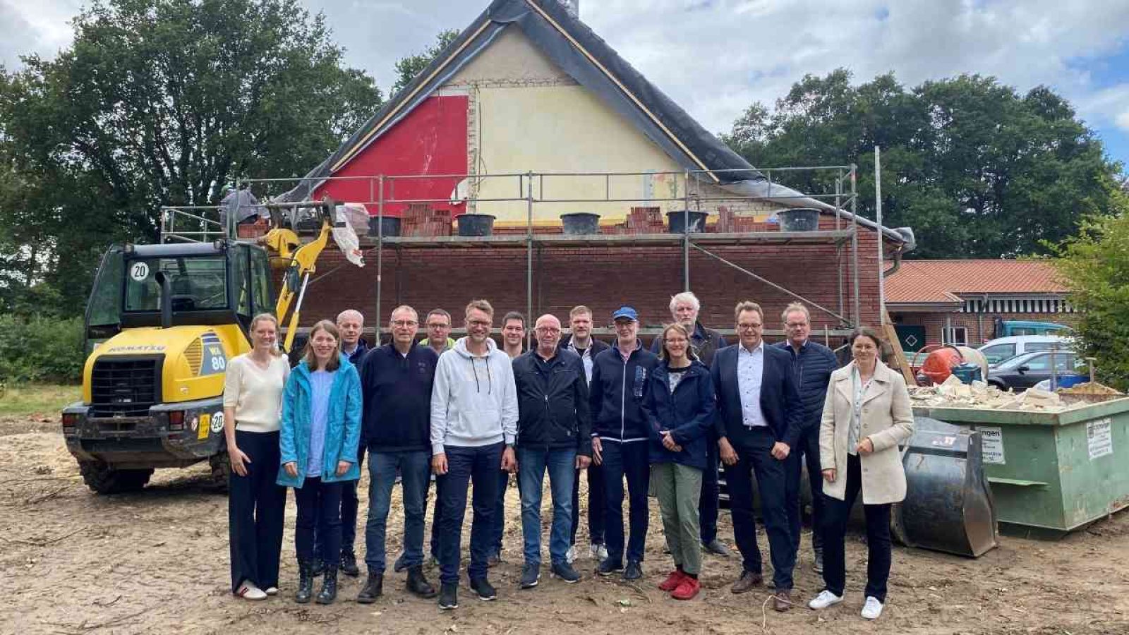 Gruppe von Menschen vor einer Baustelle mit Bagger und Hausrenovierung im Hintergrund, klarer Himmel darüber.