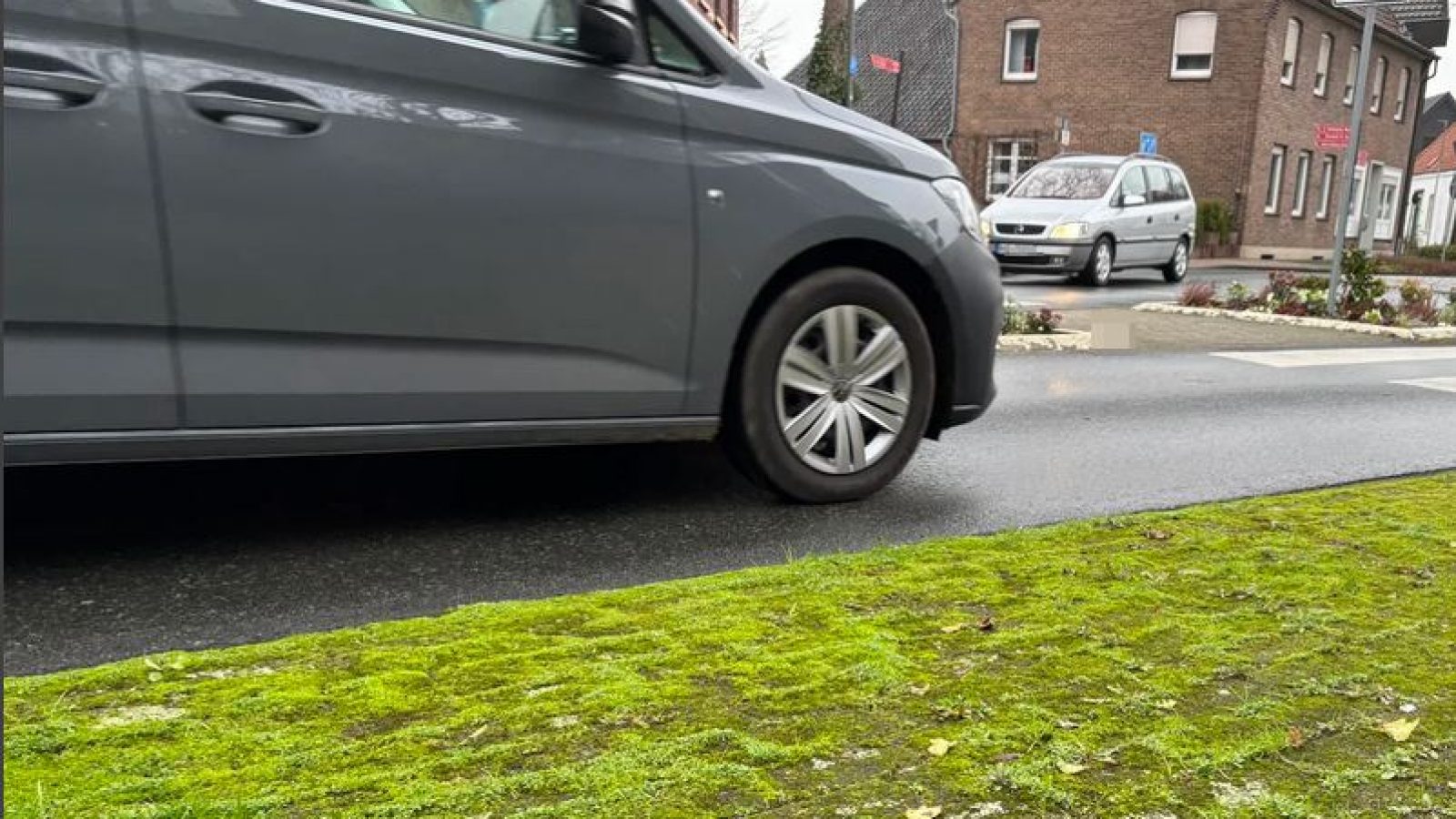 Grauer PKW, der auf einer Straße fährt, mit grünem Moos im Vordergrund.